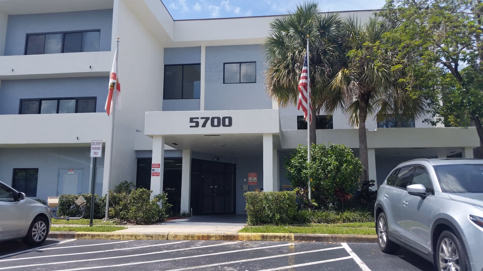Two-story building entrance with the number 5700, flags, palm trees, and parked cars.