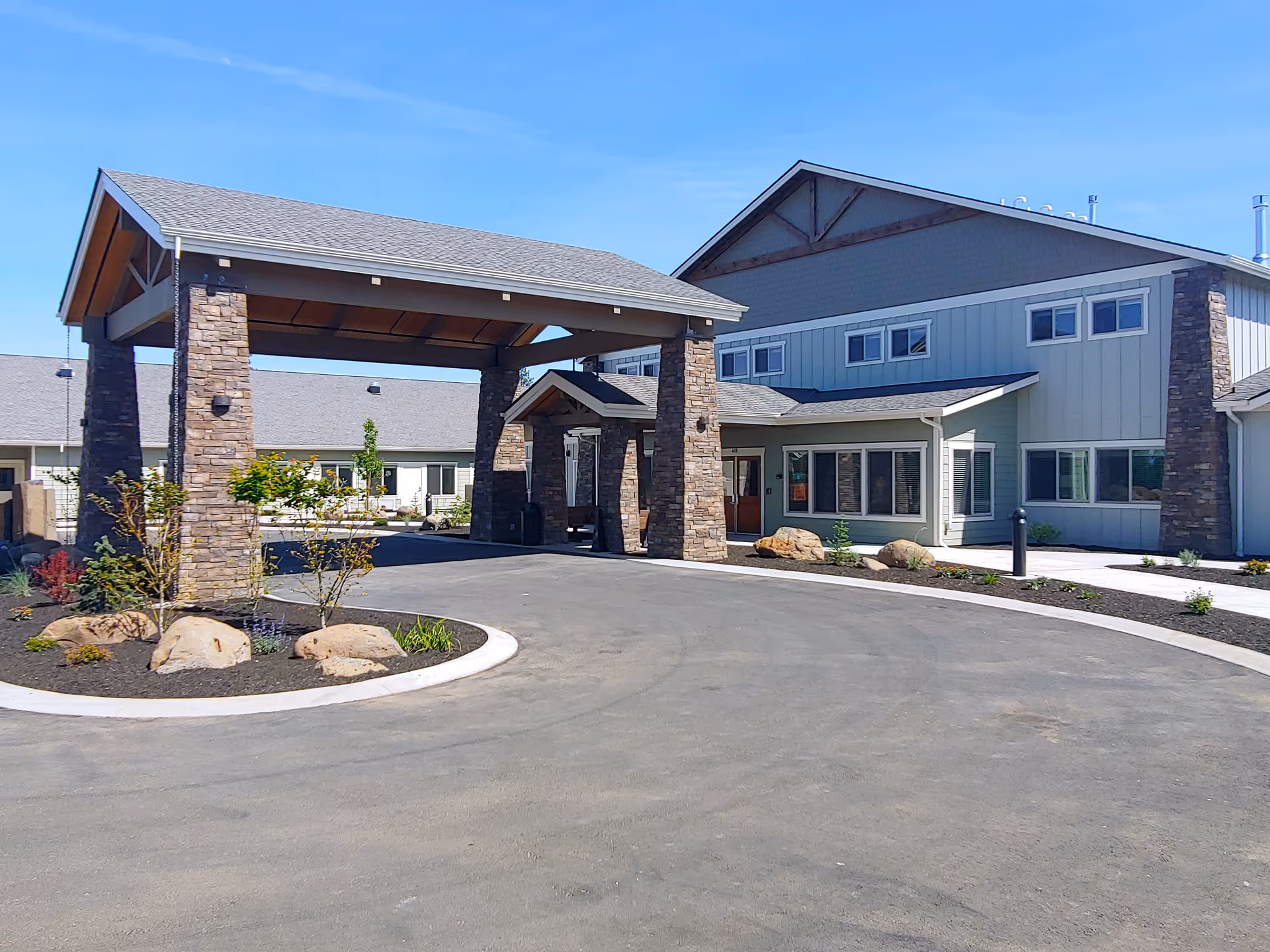 Exterior view of The Lodge Retirement Community building featuring a covered entrance with stone pillars, a paved driveway, landscaped areas with rocks and small plants, and a clear blue sky.