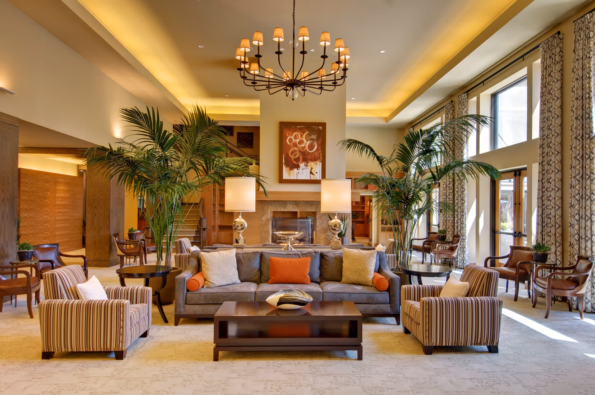 A spacious and well-lit living room area with a large gray sofa adorned with orange and cream pillows, two striped armchairs, a dark wooden coffee table with a decorative bowl, tall green plants on either side of the sofa, two table lamps with round silver bases, a modern chandelier hanging from the ceiling, large windows with patterned curtains allowing natural light, and a fireplace with abstract artwork above it.