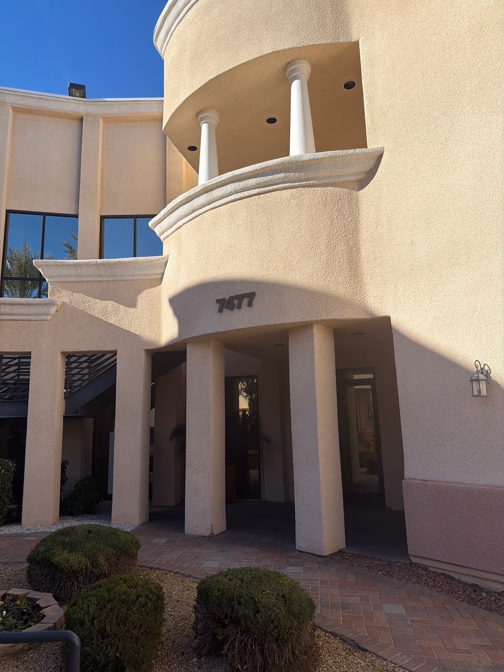Curved beige building entrance with arches and columns, address 7477, under a clear blue sky.