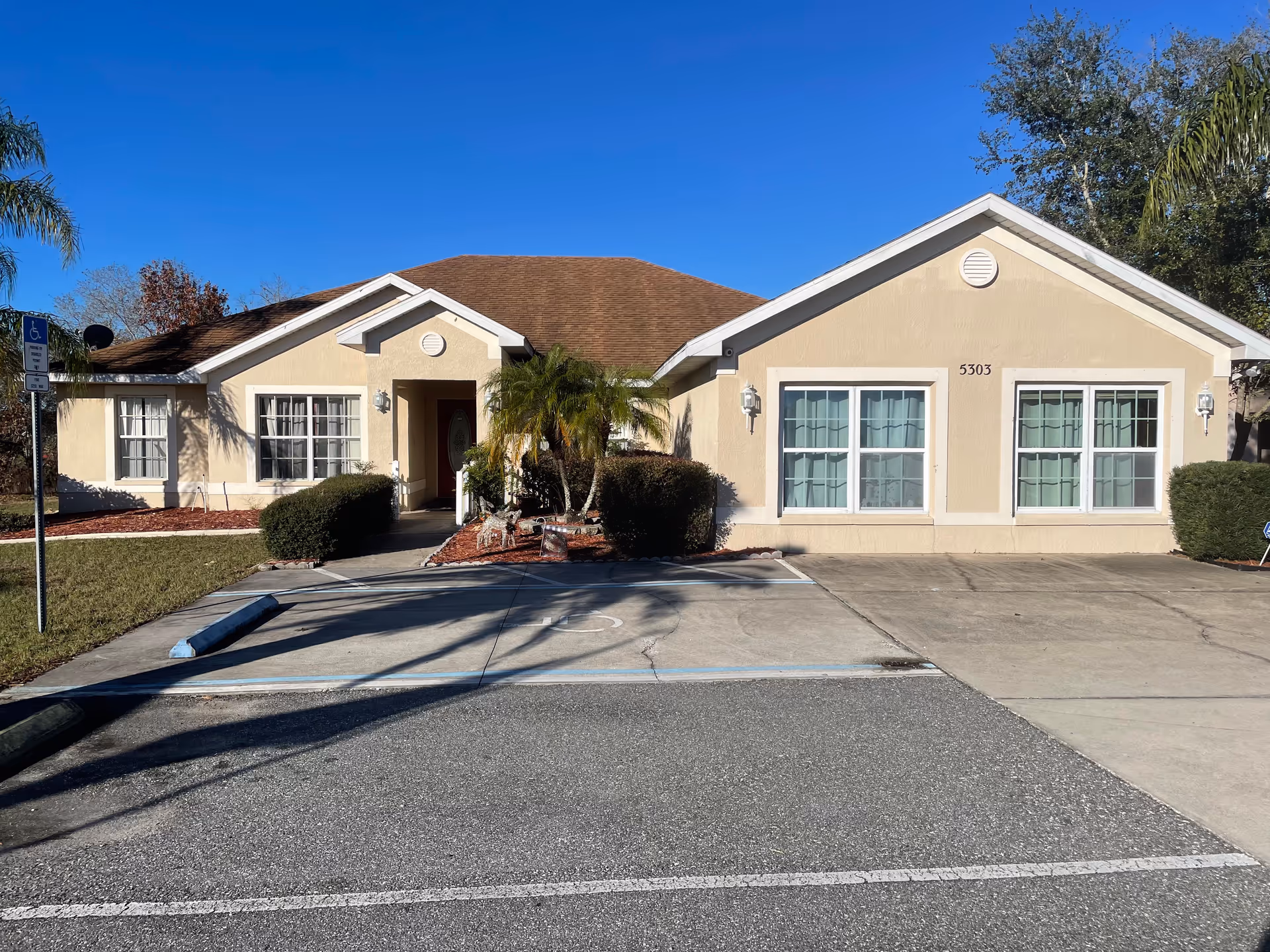 Exterior front view of a single-story beige building with a brown roof, featuring multiple windows with white frames and a central entrance surrounded by bushes and palm plants. There is a handicapped parking space in front of the building under a clear blue sky.
