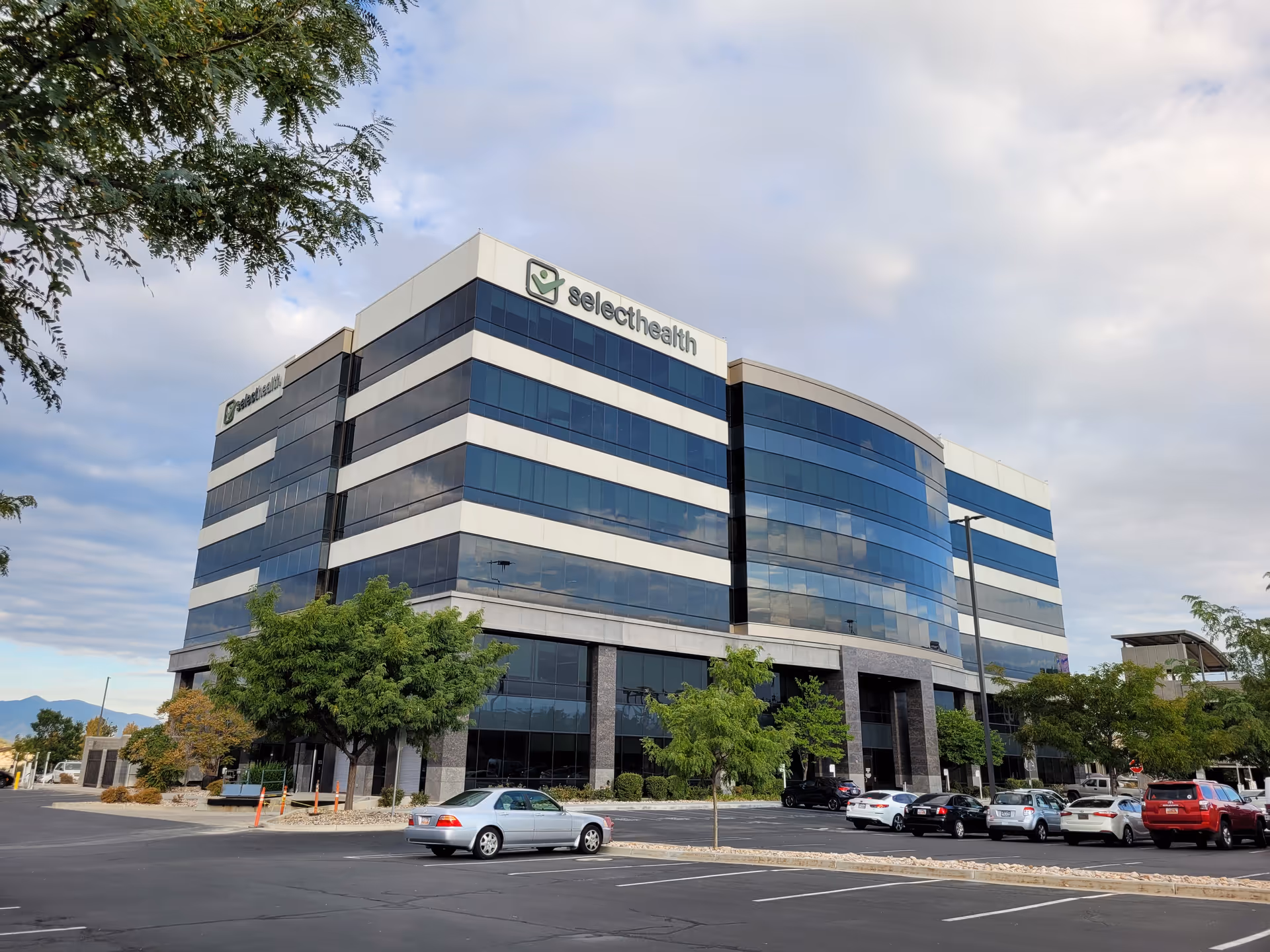 Modern Select Health building with glass facade, parking lot, and surrounding trees.