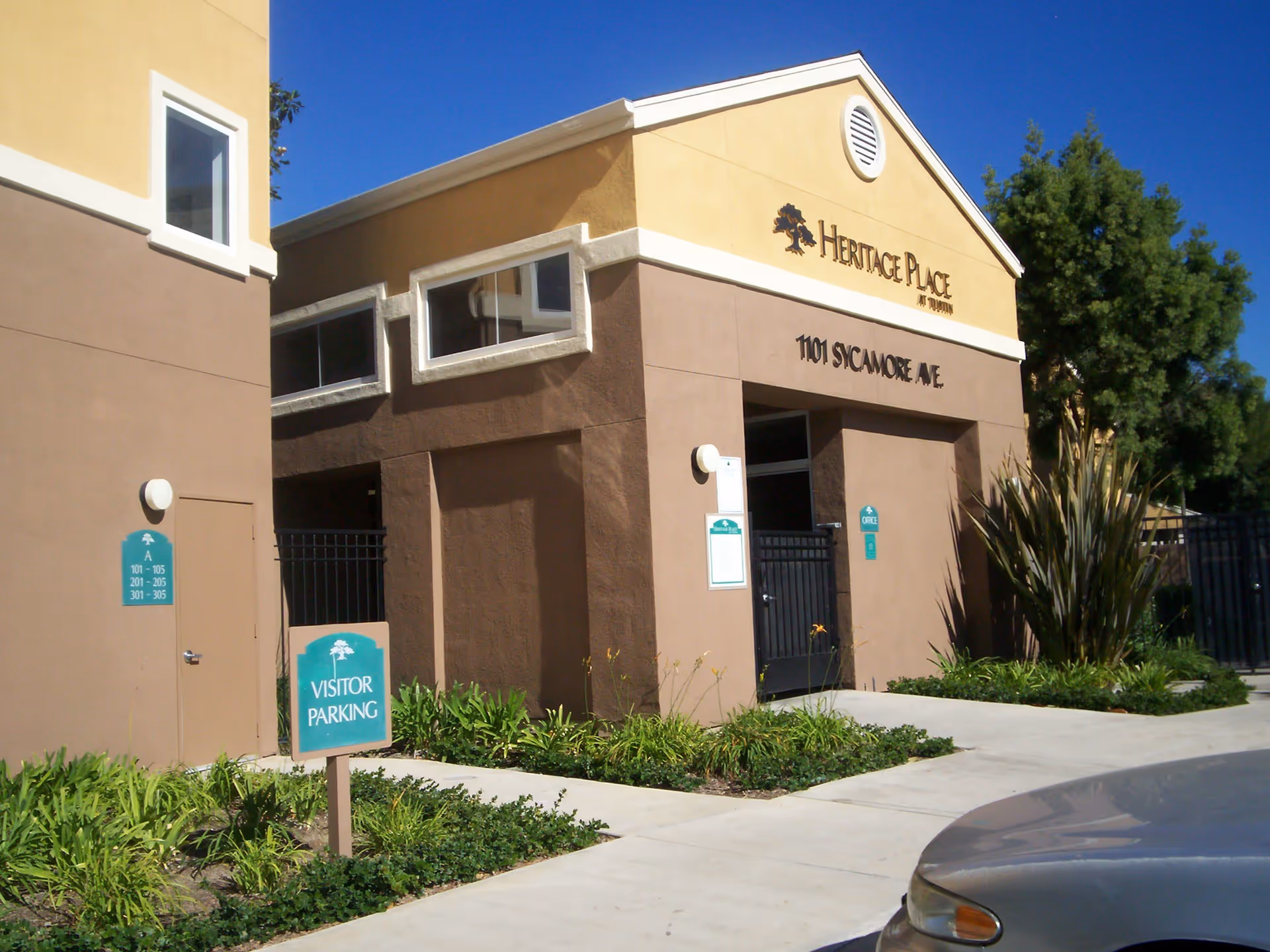 Exterior front entrance of the Heritage Place at Tustin building with a 'Visitor Parking' sign and surrounding landscaping.