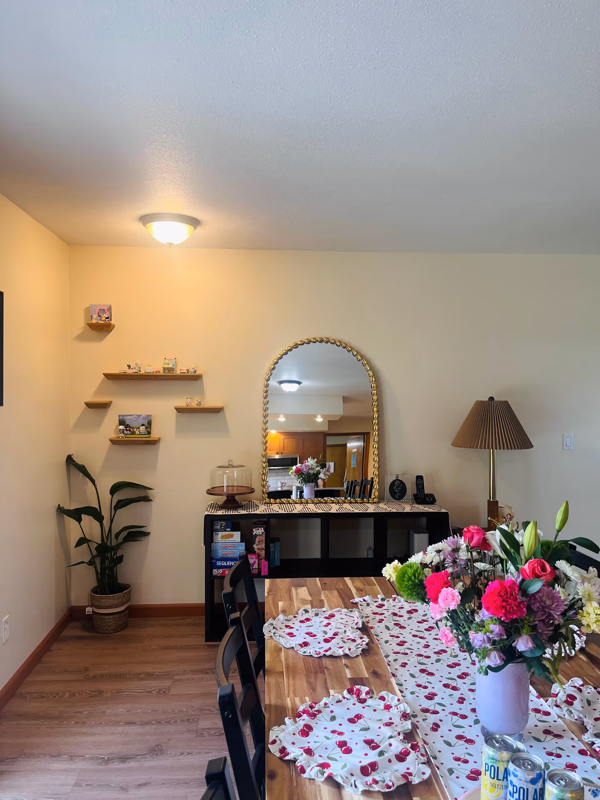 Dining room scene with a wooden table, floral centerpiece, shelves, and mirror.