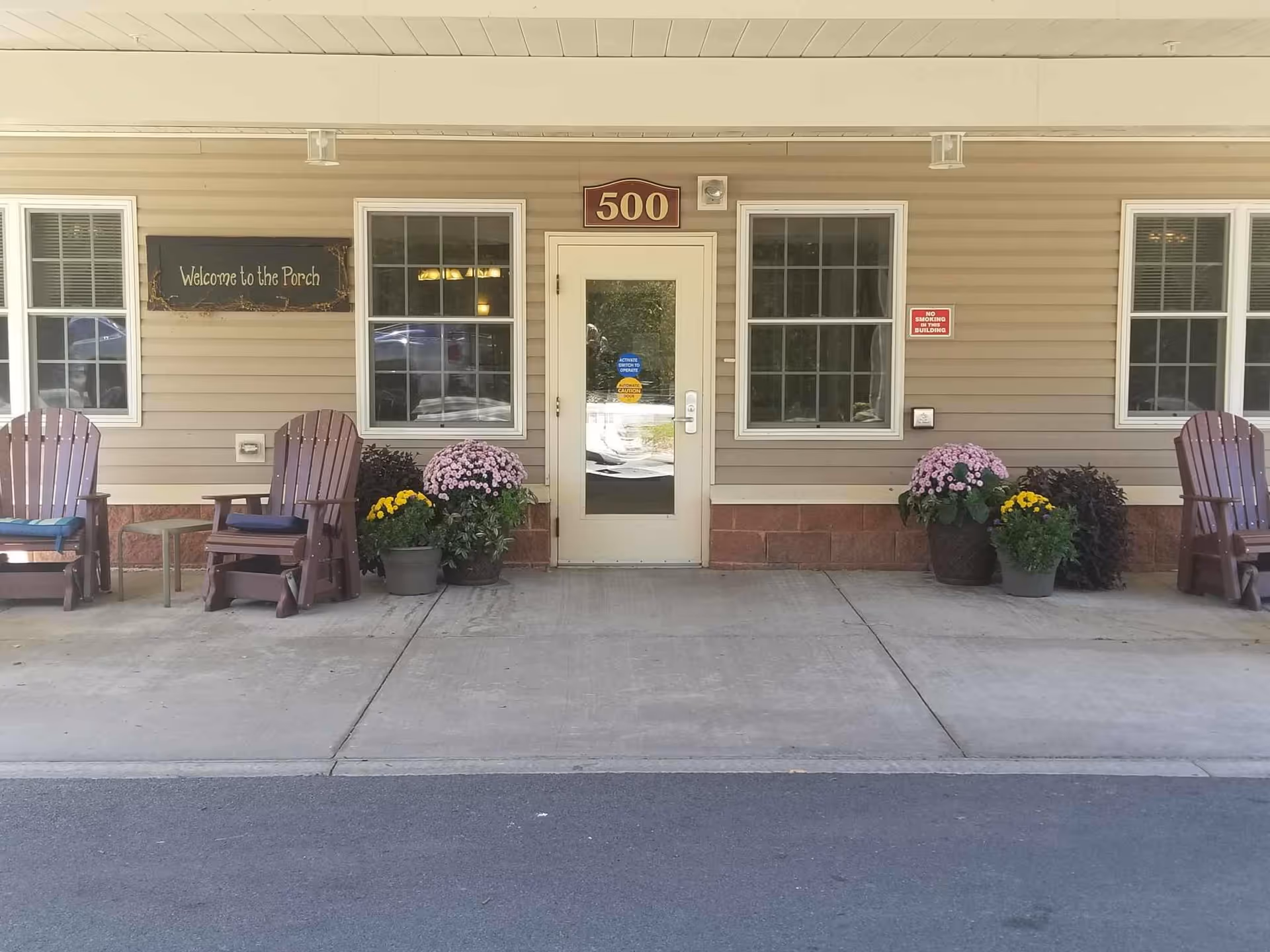 Front entrance of a building with the number 500 above the door. The entrance has a glass door flanked by two windows on each side. There are four wooden chairs with cushions and several potted plants with flowers arranged on the porch. A sign on the left side reads 'Welcome to the Porch' and a small red sign on the right side says 'No Smoking Building'.