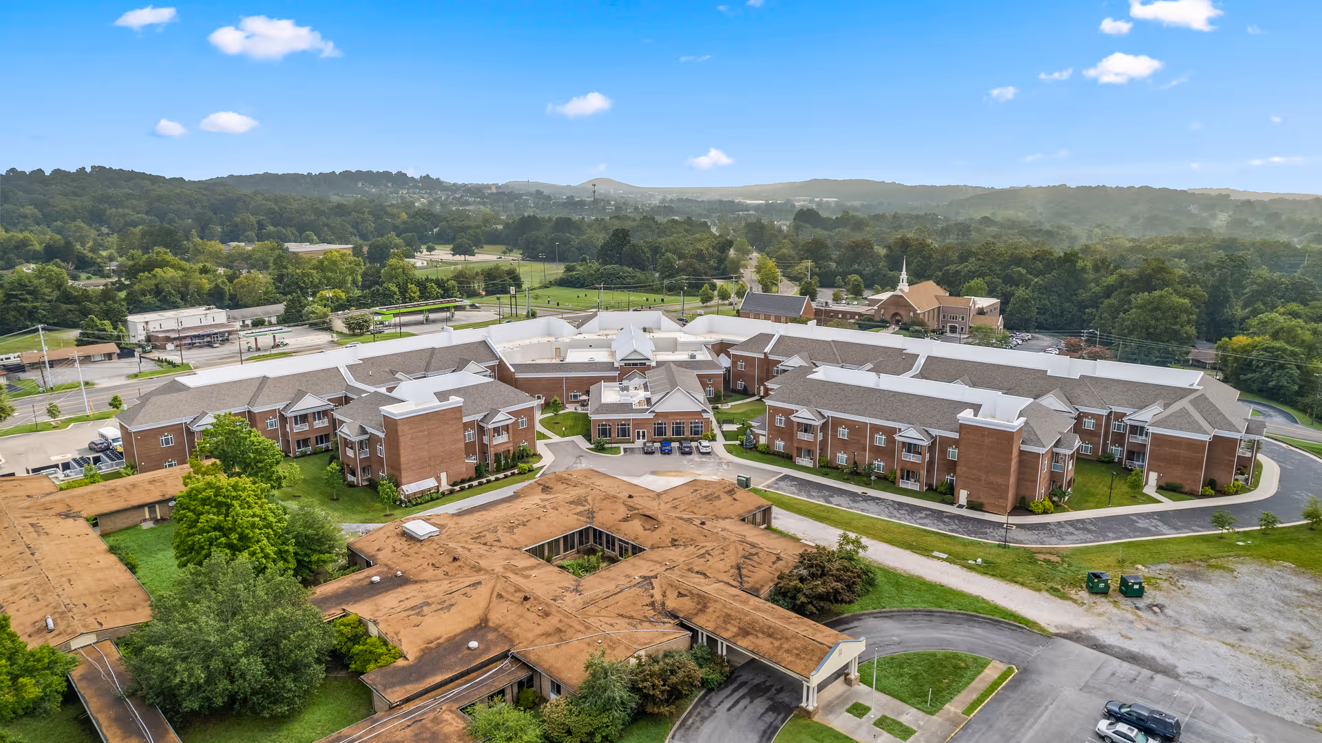 Aerial view of Shannondale of Knoxville senior living facility showing multiple connected brick buildings with gray roofs, surrounded by green trees and lawns, with a road and parking areas nearby under a blue sky with scattered clouds.