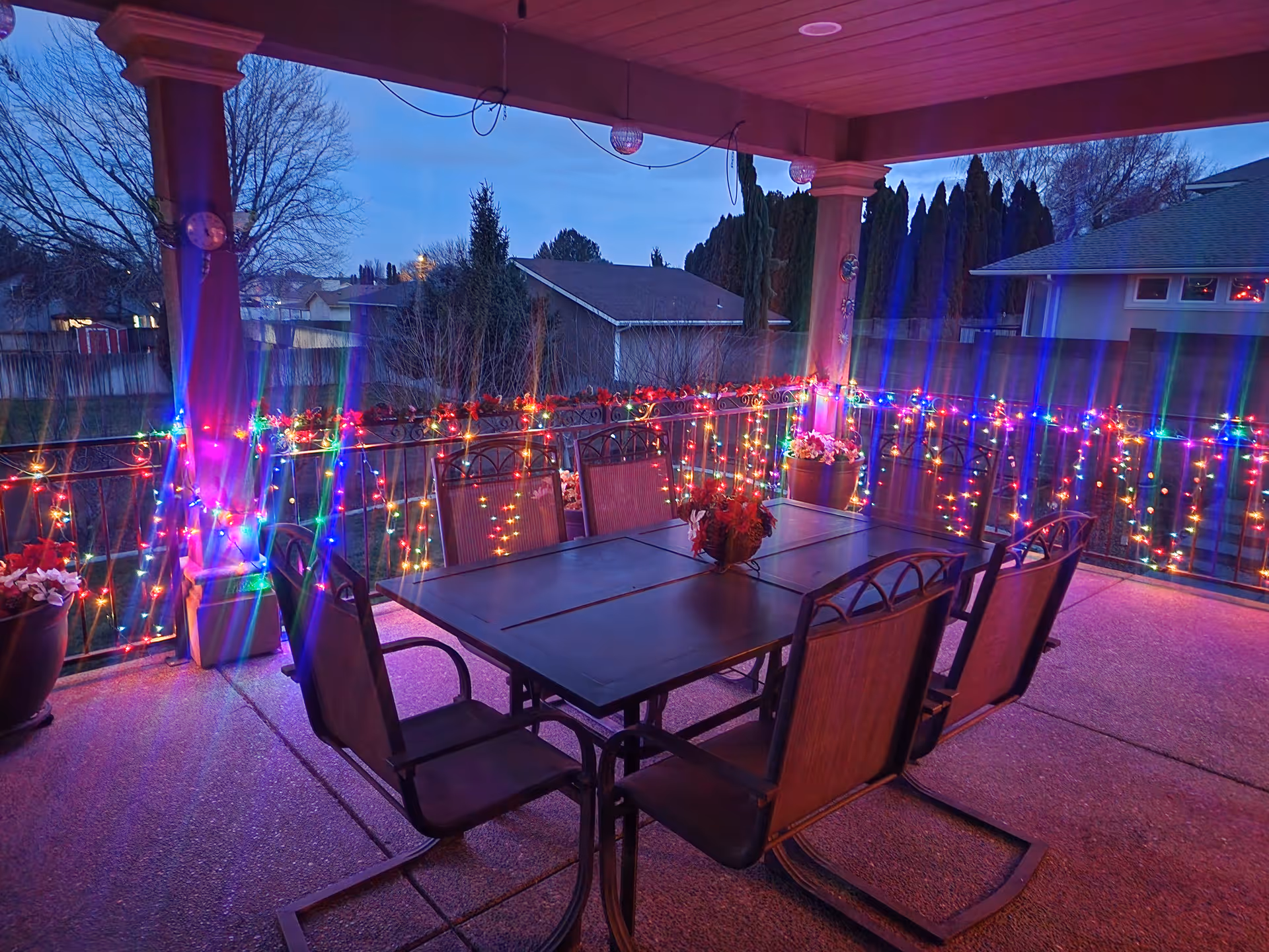 Covered outdoor patio area at dusk with a rectangular dining table and six chairs. The railing around the patio is decorated with colorful string lights, and there are potted plants with flowers placed near the corners. Trees and neighboring houses are visible in the background.