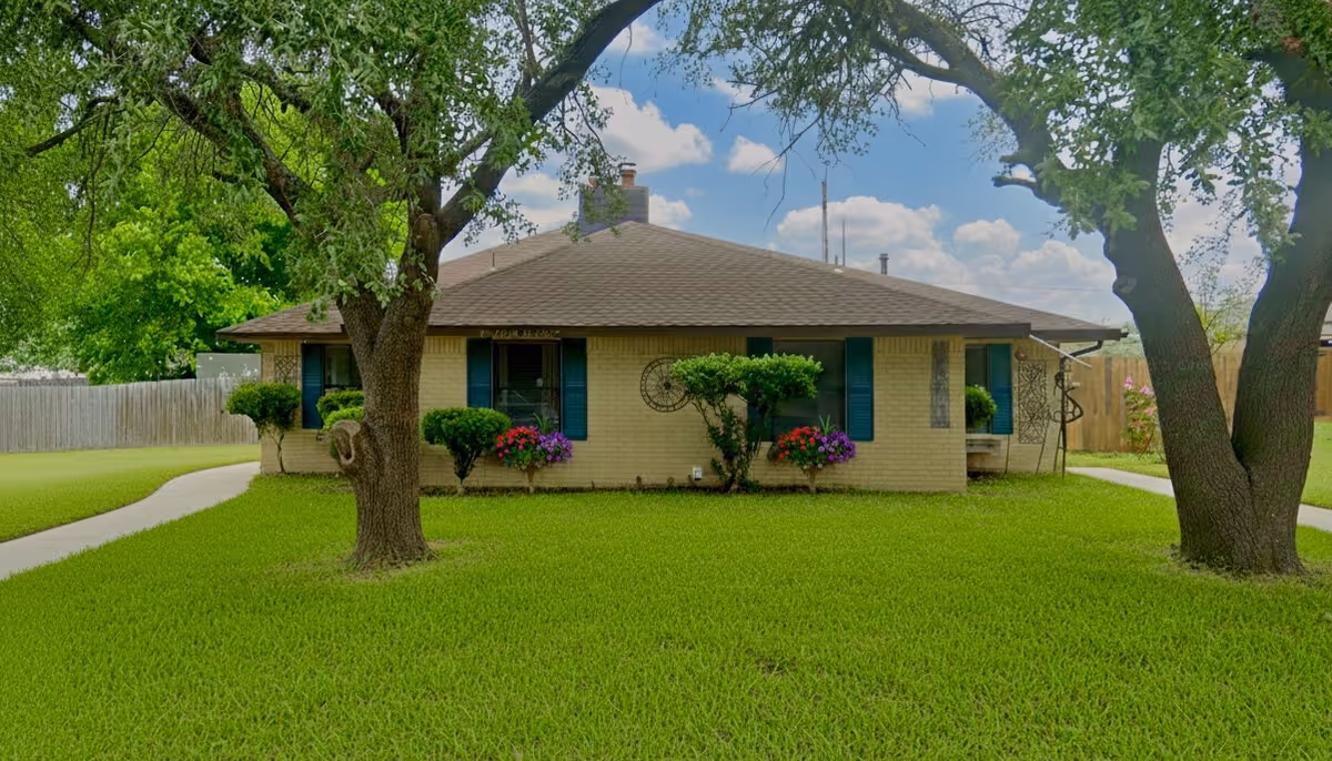 Single-story beige brick senior living home with green lawn and large trees in front.
