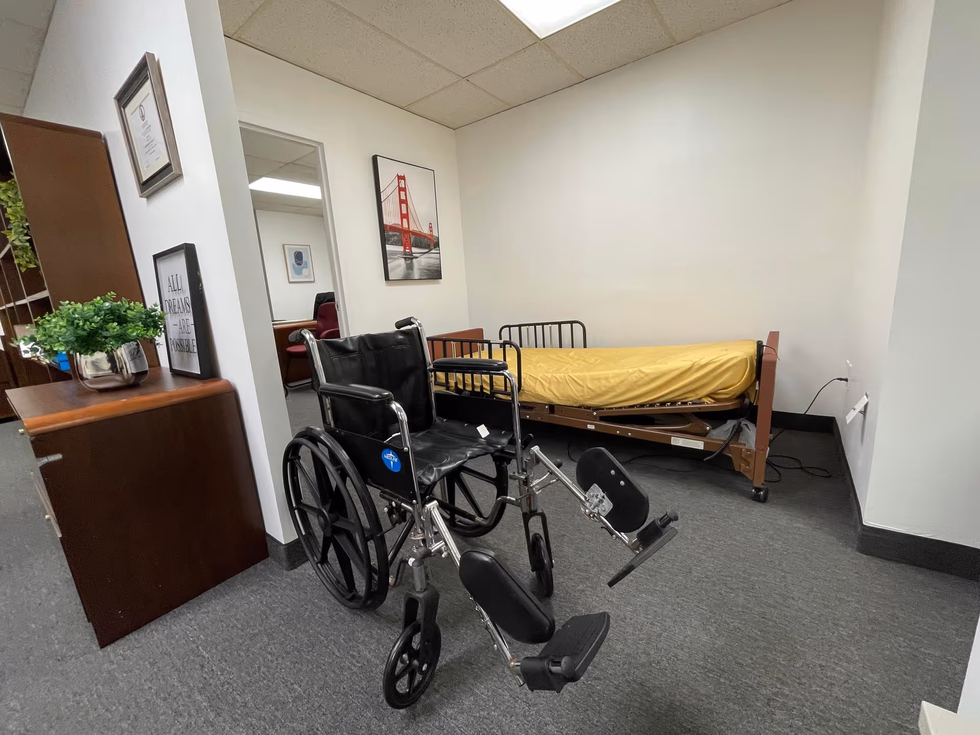 Wheelchair and hospital bed in a small care room with framed pictures and a plant.