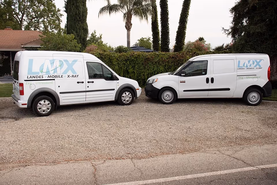 Two white Landes Mobile X-ray vans parked on a gravel lot with greenery behind.