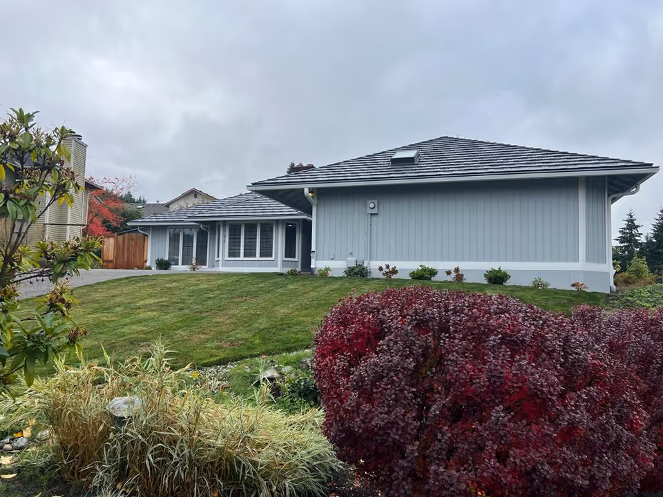 Front exterior of a single-story light-blue house with a manicured lawn, shrubs, and an overcast sky.