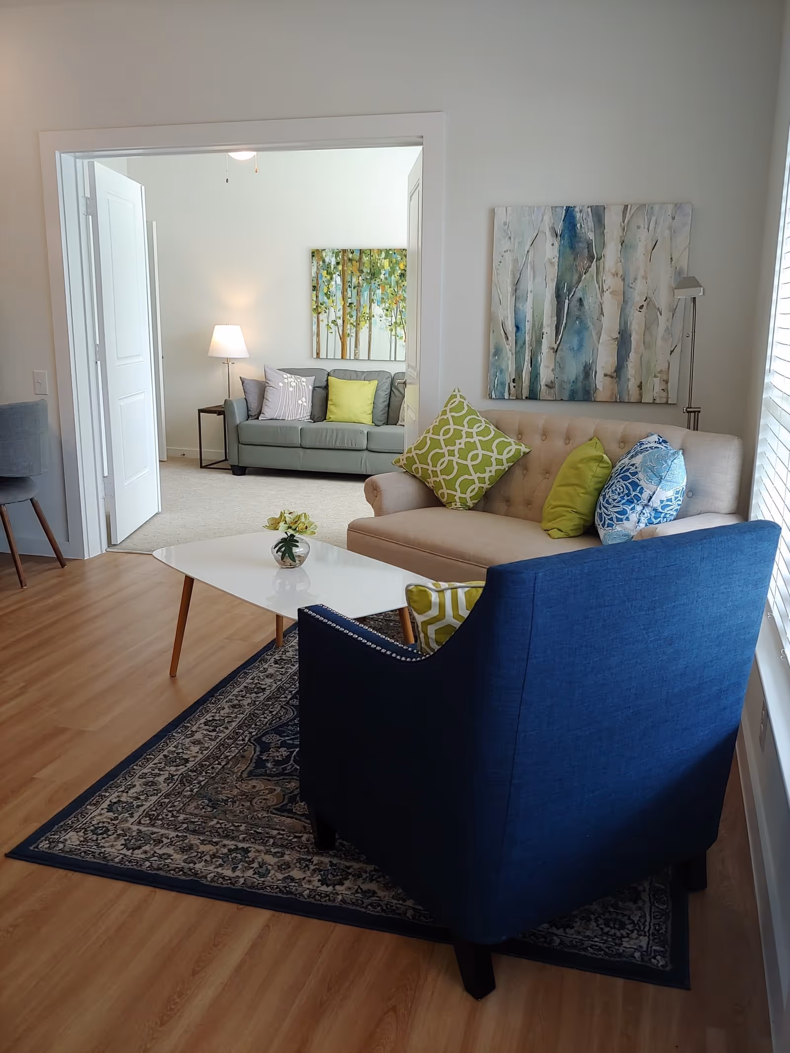A cozy living room area with a beige tufted sofa adorned with green and blue patterned pillows, a blue armchair, and a white coffee table with a small flower vase. The room has wooden flooring and a patterned rug. In the background, through an open doorway, there is another seating area with a gray sofa, colorful pillows, a side table, and a lamp. Both rooms have light-colored walls with nature-themed artwork.
