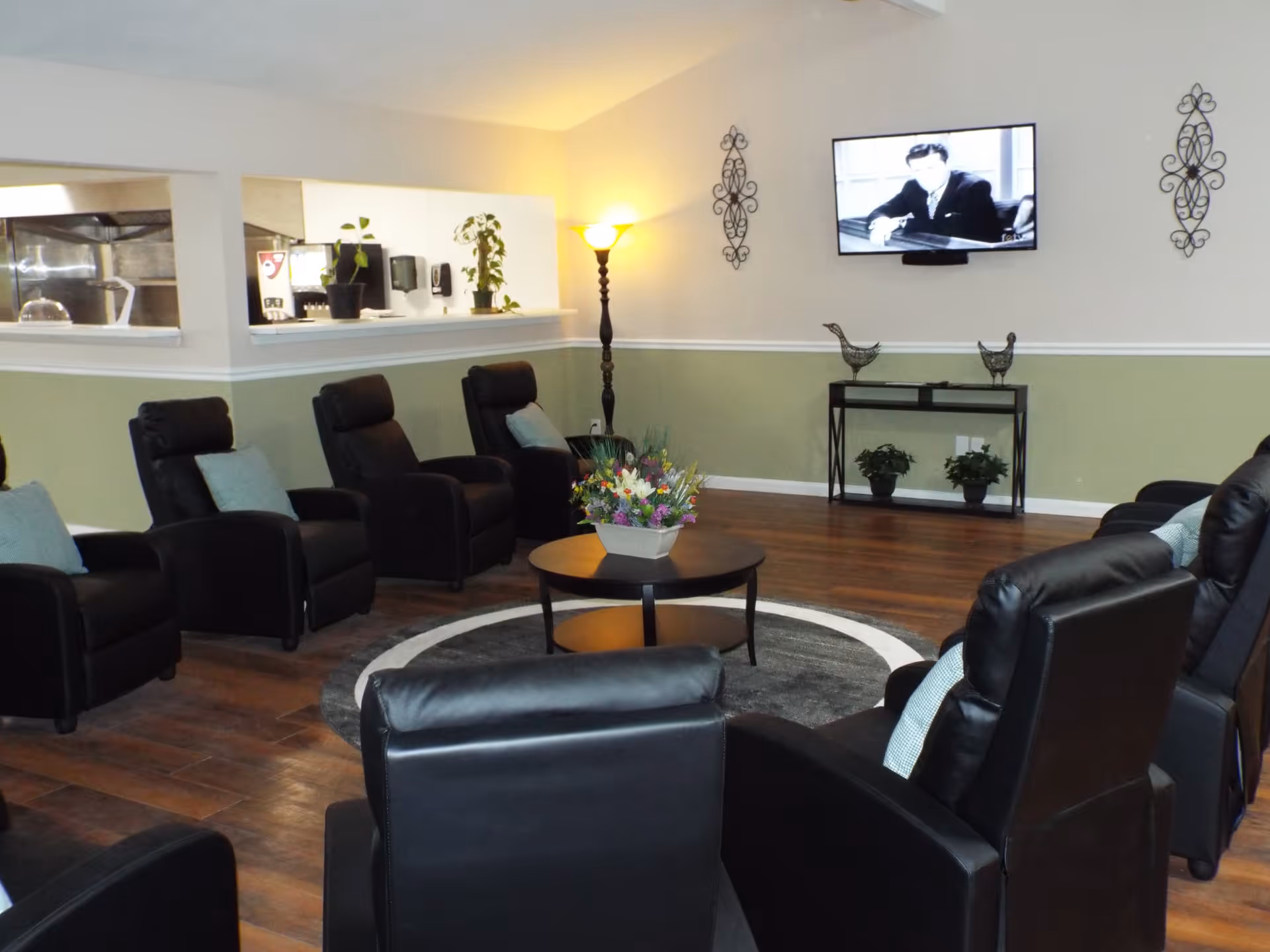 A cozy living room area with black leather recliners arranged in a semi-circle around a round coffee table with a flower arrangement. The room has wooden flooring, a green and white painted wall, a tall floor lamp, and a wall-mounted TV showing a black and white image. There are decorative wall hangings and plants on a small table beneath the TV.