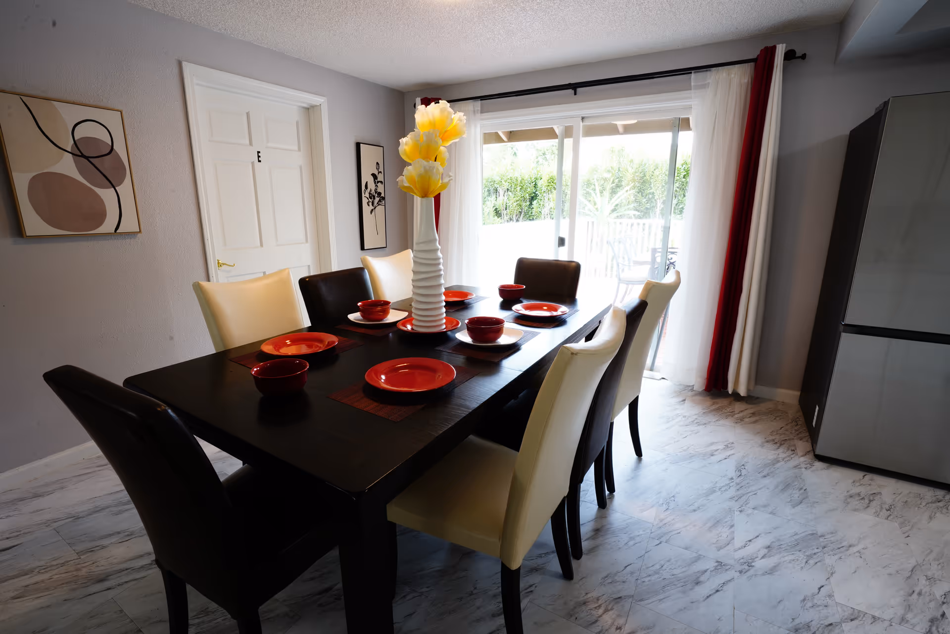 A dark dining table with eight chairs and red dishes in a bright dining room.