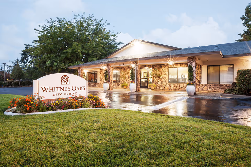 Whitney Oaks care center exterior with stone facade, illuminated entry, and landscaped front lawn.