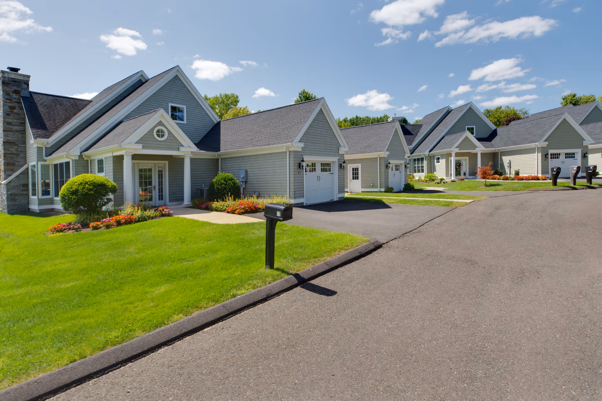 Exterior view of a senior living facility with multiple attached single-story homes featuring gray siding, white trim, garages, well-maintained green lawns, flower beds, and a paved road under a partly cloudy blue sky.