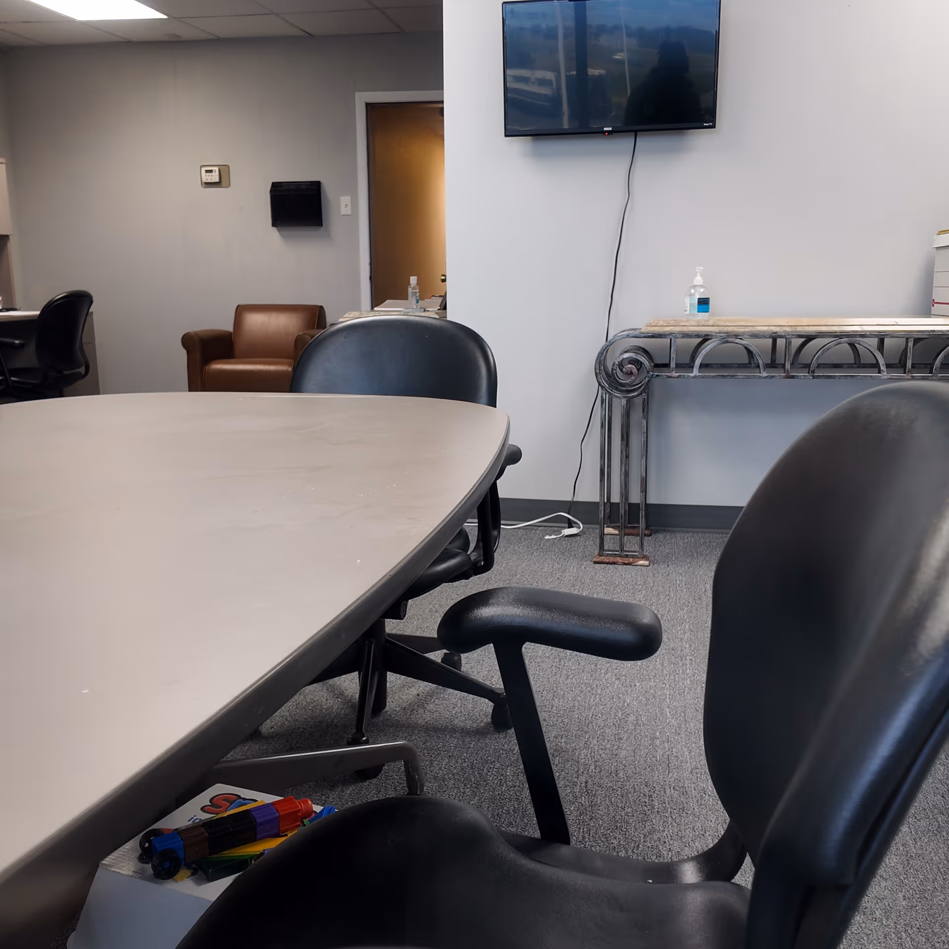 Empty conference room with large table, chairs, wall-mounted TV, and sanitizer bottle.