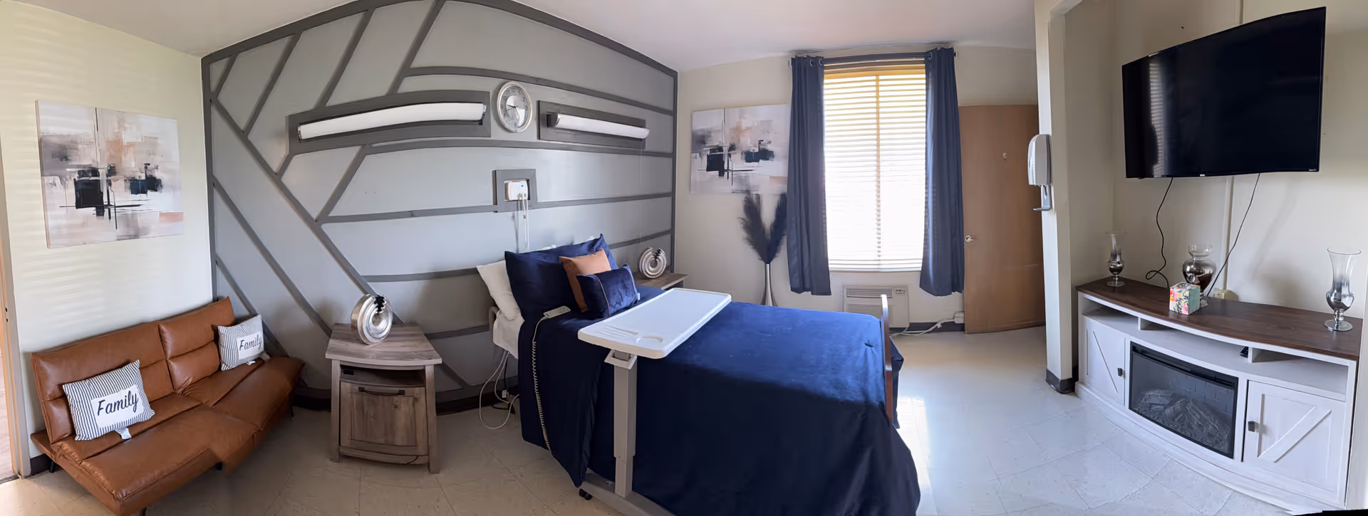 Blue-blanket bed in a modern senior room with a geometric gray wall, sofa, and mounted TV.