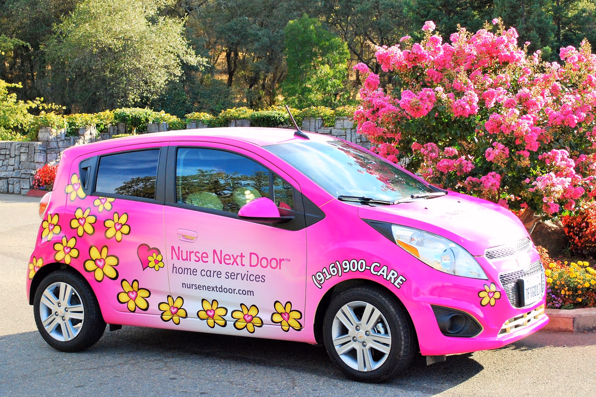 Bright pink Nurse Next Door van with yellow flowers parked outdoors beside blooming bushes.