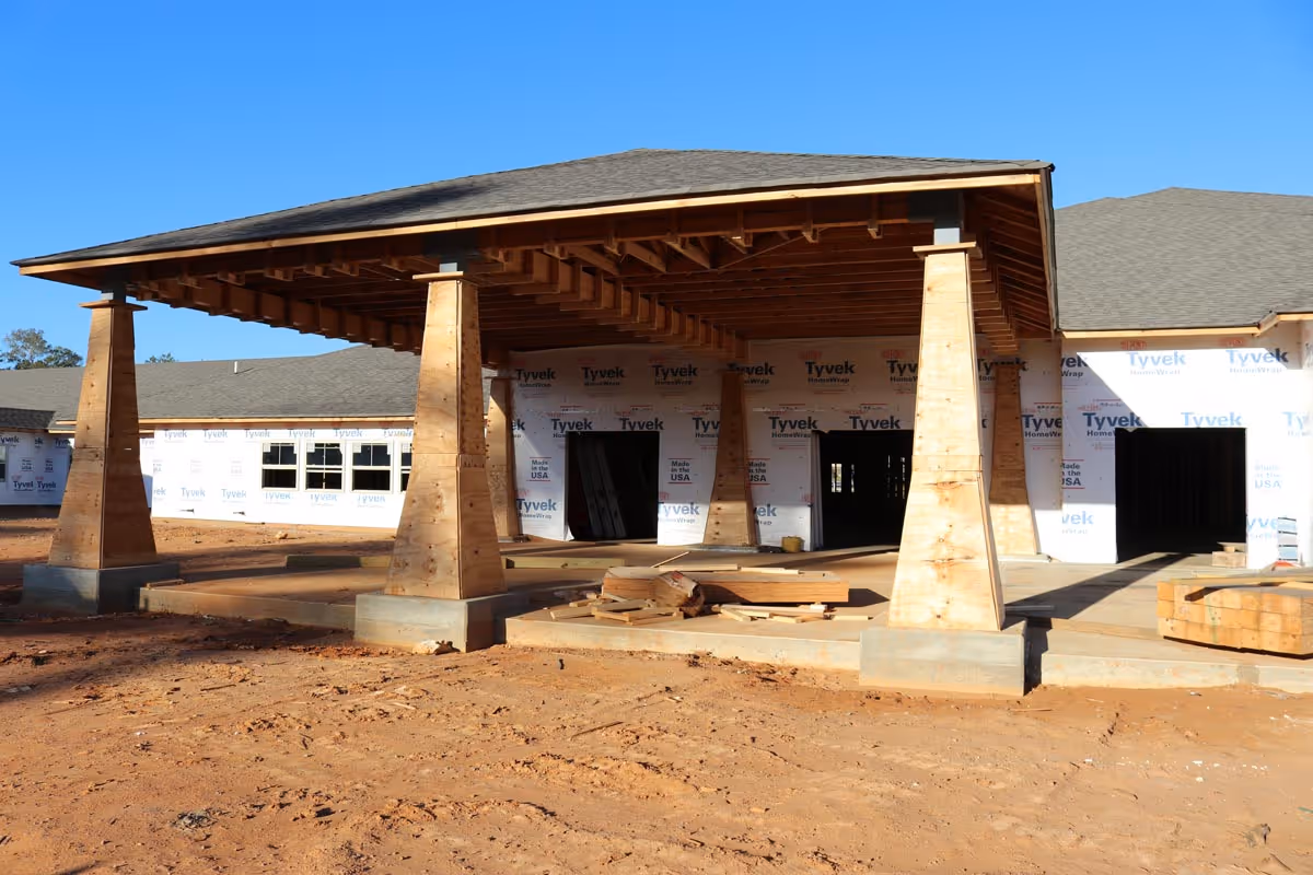 A building under construction with a large covered porch supported by wooden pillars. The exterior walls are wrapped in Tyvek HomeWrap, and construction materials are scattered on the ground. The sky is clear and blue.