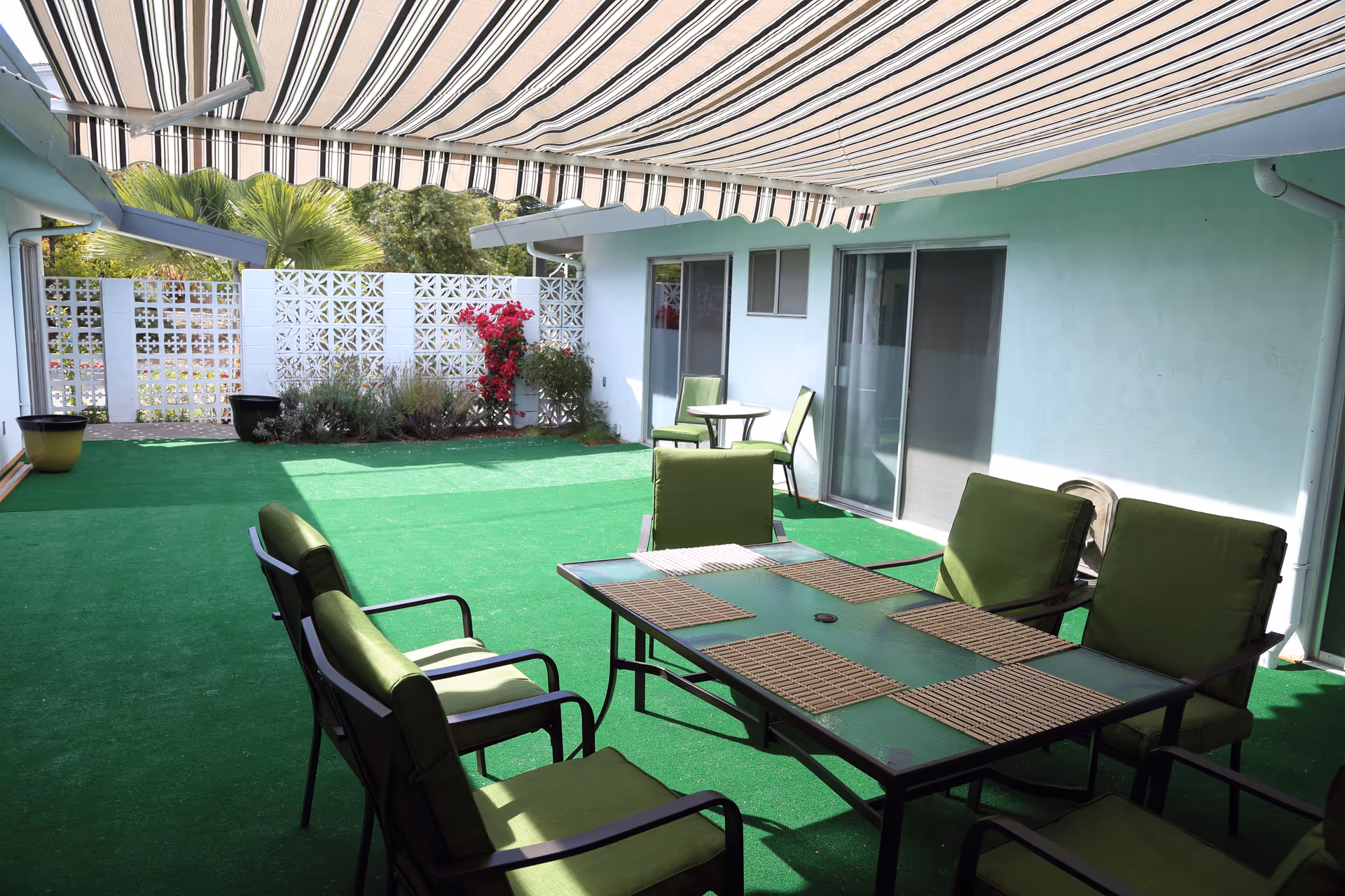Outdoor patio area with green cushioned chairs around a glass table with placemats, a smaller table with two chairs in the background, green artificial turf flooring, and a striped retractable awning overhead. There are plants and flowers along a white lattice wall.