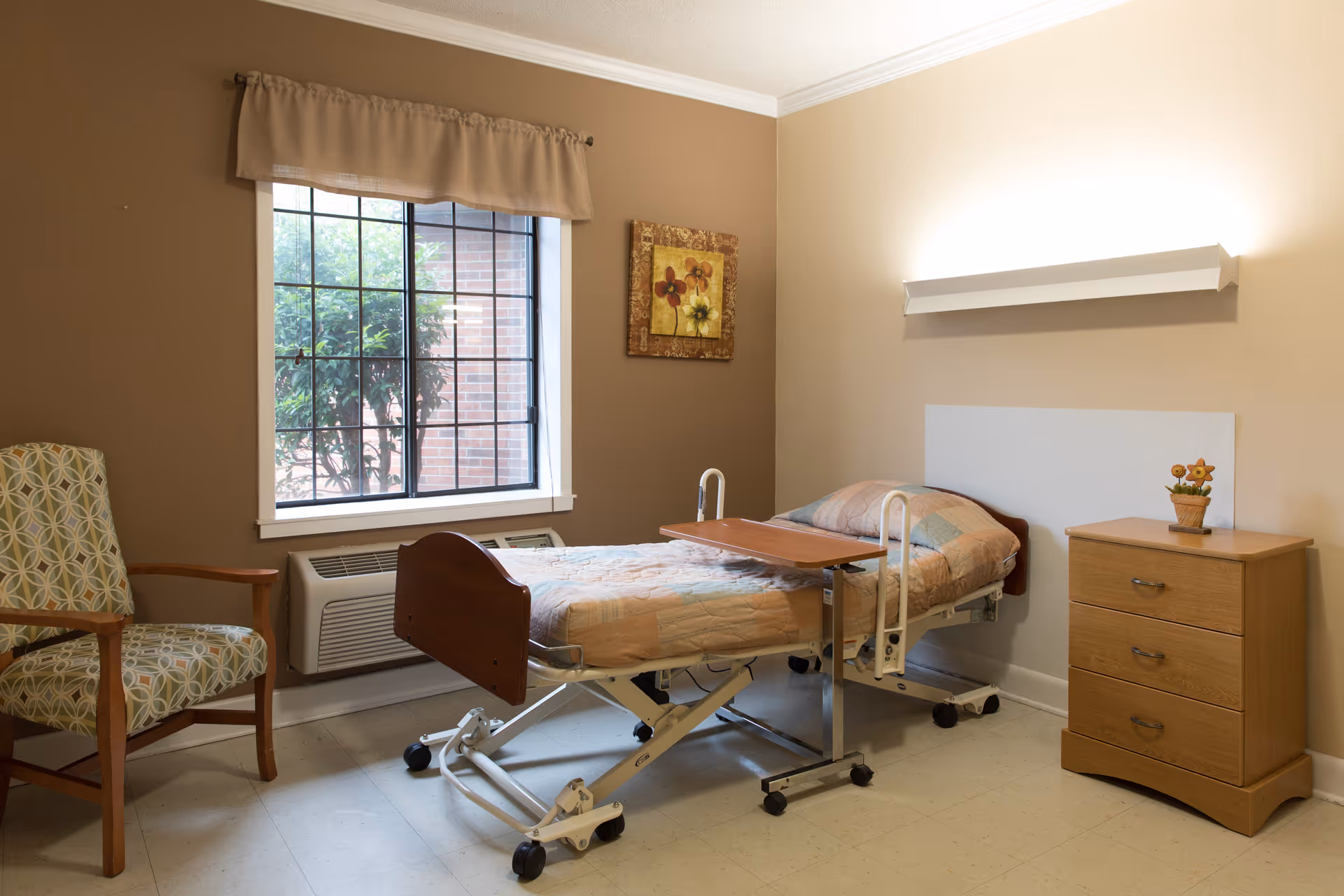 A small, neatly arranged room with a single hospital-style bed covered with a patchwork quilt. Next to the bed is a wooden bedside table with three drawers and a small flower pot on top. There is a patterned armchair near a window with a beige valance, and a wall-mounted light fixture above the bed. The walls are painted in two tones of beige and brown.