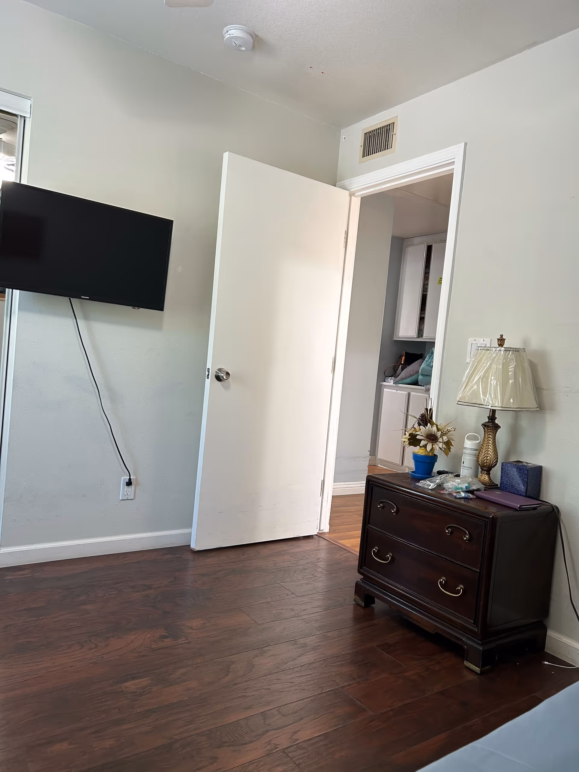 Small bedroom with hardwood floors, a wall-mounted TV, an open door leading to a hallway, and a dark wood nightstand topped with a lamp and flowers.