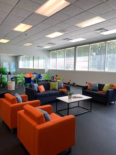 Bright, modern senior living lounge with colorful couches, tables, and large windows.