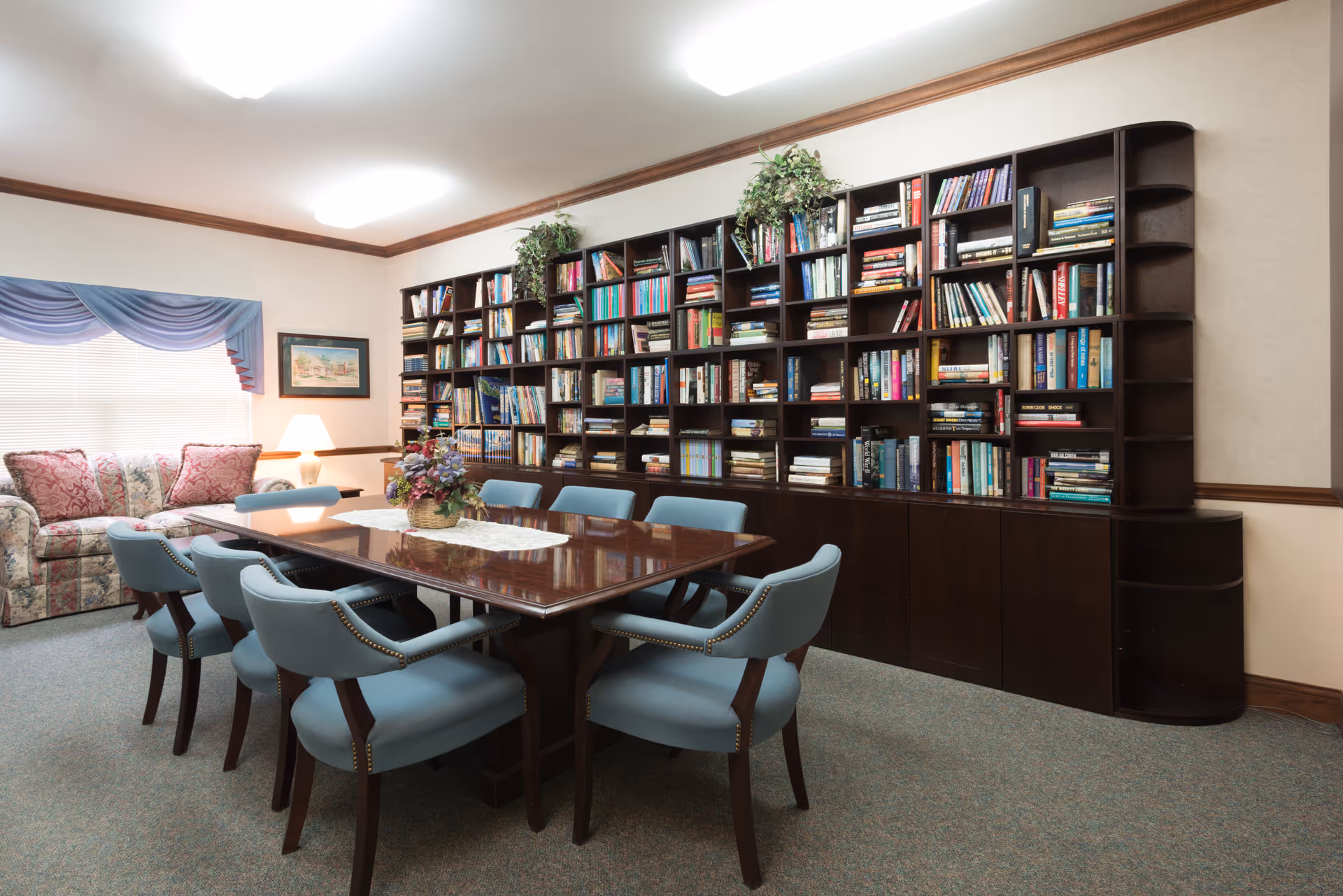 A cozy room with a large wooden table surrounded by eight blue upholstered chairs. Behind the table is a tall bookshelf filled with numerous books and some decorative plants. To the left, there is a floral-patterned sofa with two pink cushions, a window with blue drapes, and a table lamp on a side table. The room has carpeted flooring and soft overhead lighting.
