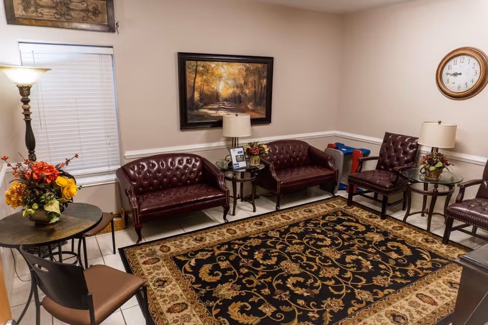 A waiting lounge with burgundy tufted chairs, glass tables, a patterned rug, and a wall clock.