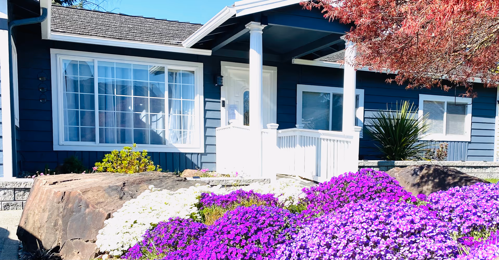Blue single-story senior living facility exterior with white porch and colorful flowers.