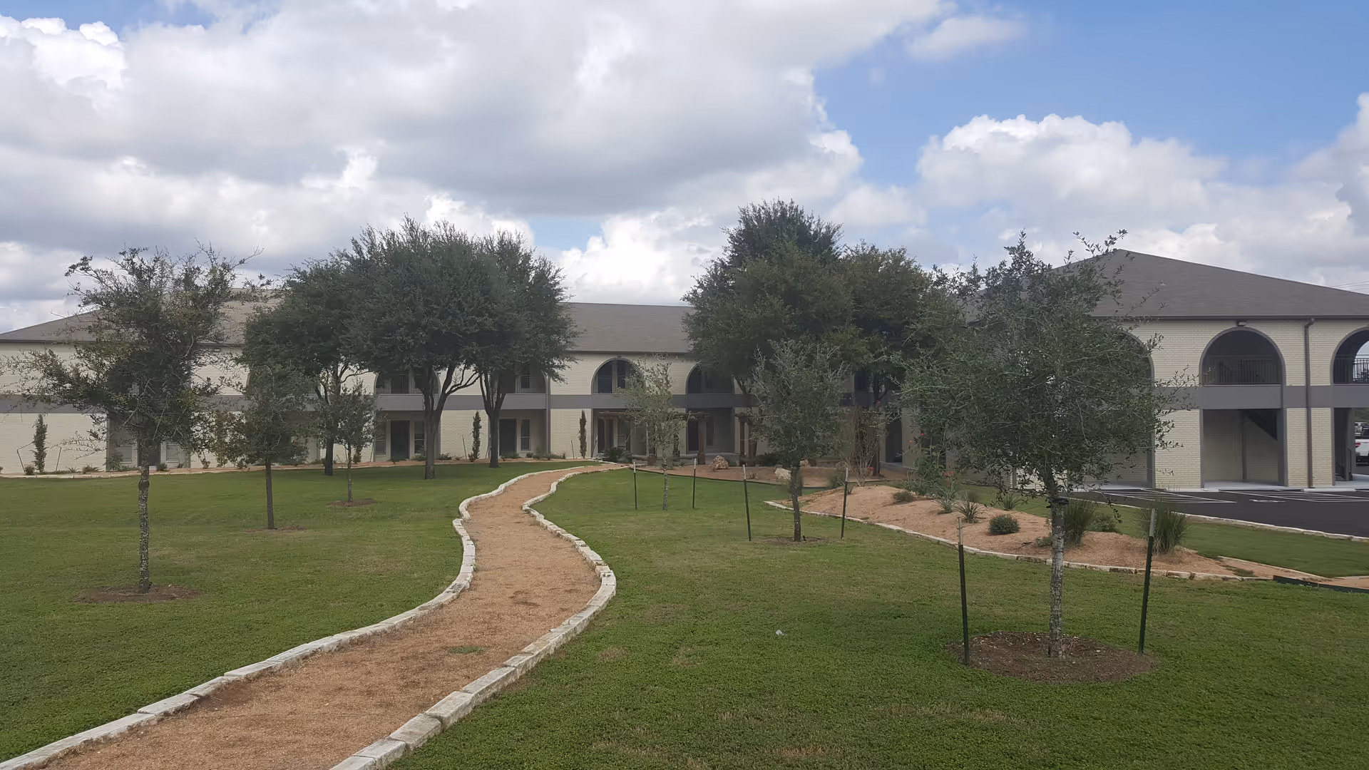 Exterior view of a senior living facility with a winding path, lawn, and trees.