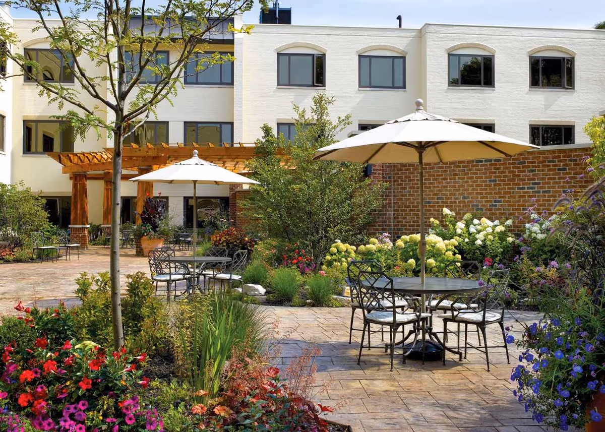 Outdoor patio area at a senior living facility with metal tables and chairs under large beige umbrellas, surrounded by colorful flowers, shrubs, and trees. The background shows a white multi-story building with large windows and a wooden pergola.