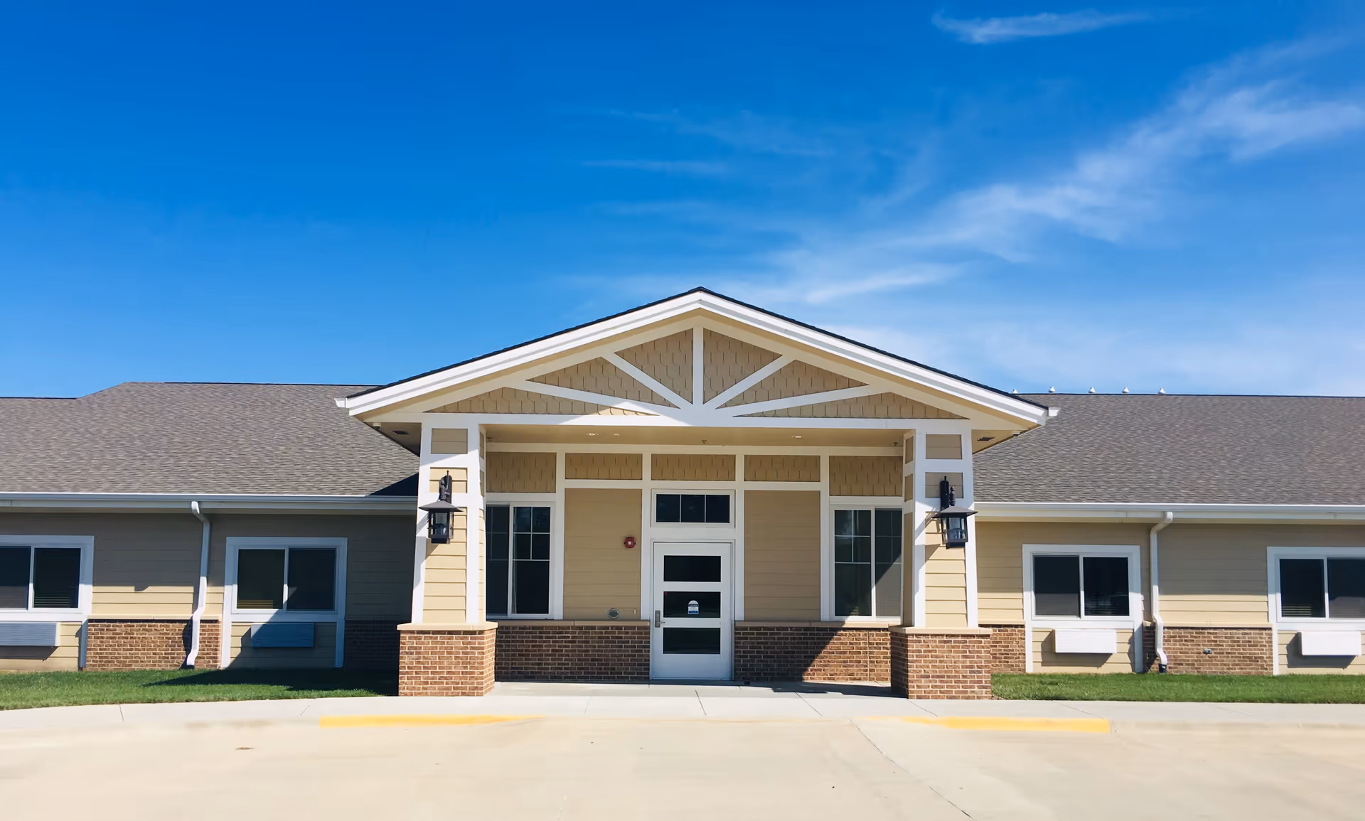 Front exterior view of Hansen House, a single-story building with beige siding, brick accents, and a covered entrance under a gabled roof. The sky is clear and blue.