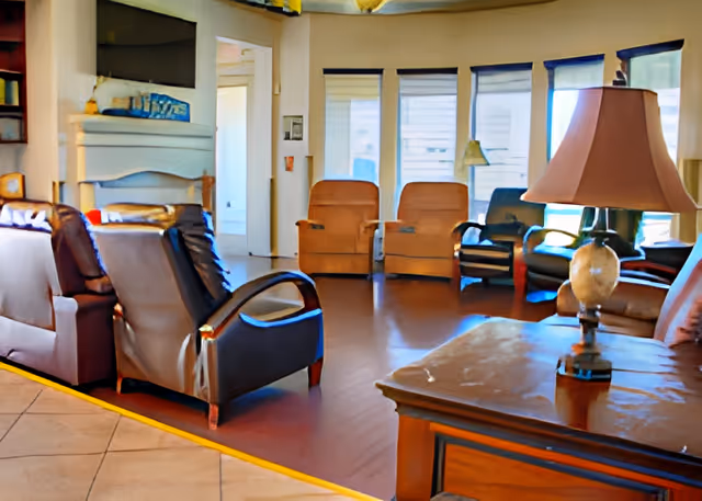 Sunlit common living room with multiple lounge chairs, a side table with a lamp, and large windows.