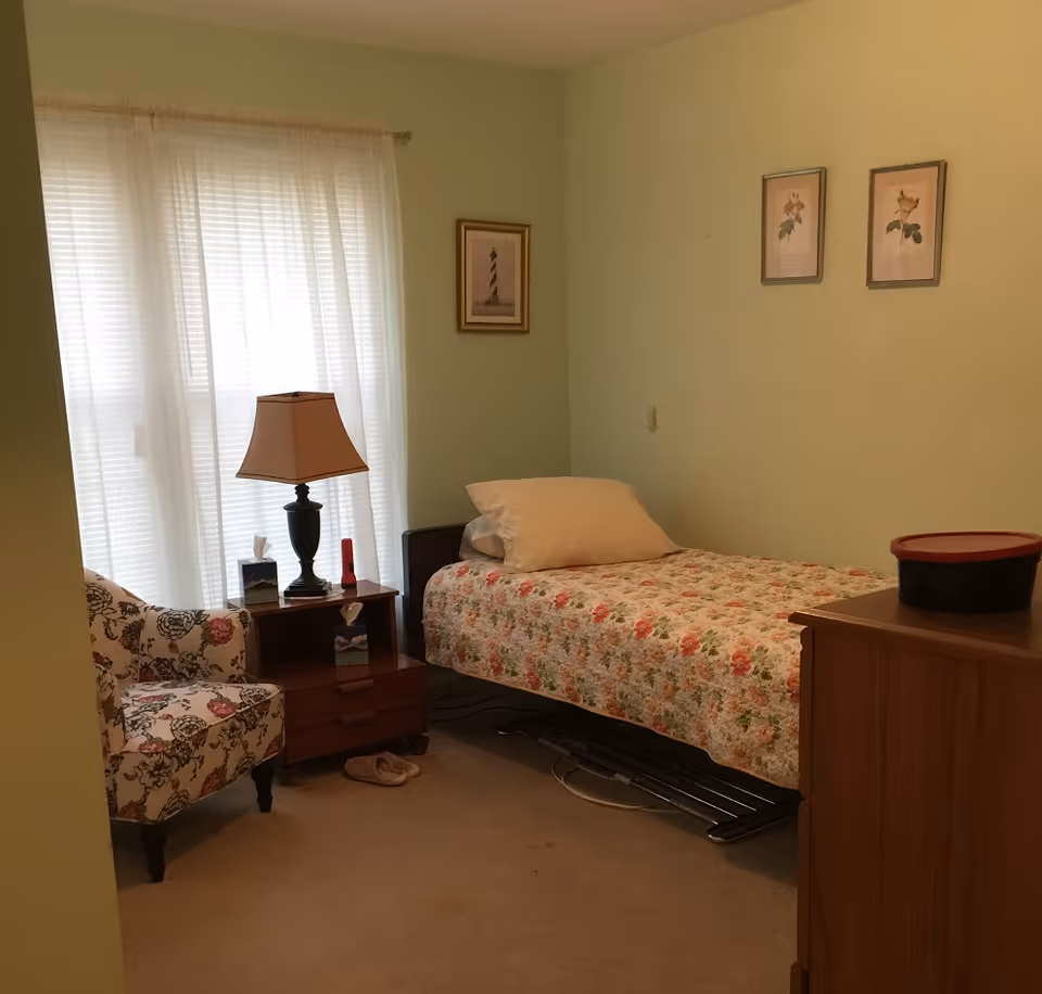 Pastel-green bedroom with single bed, floral quilt, nightstand, lamp, and chair.