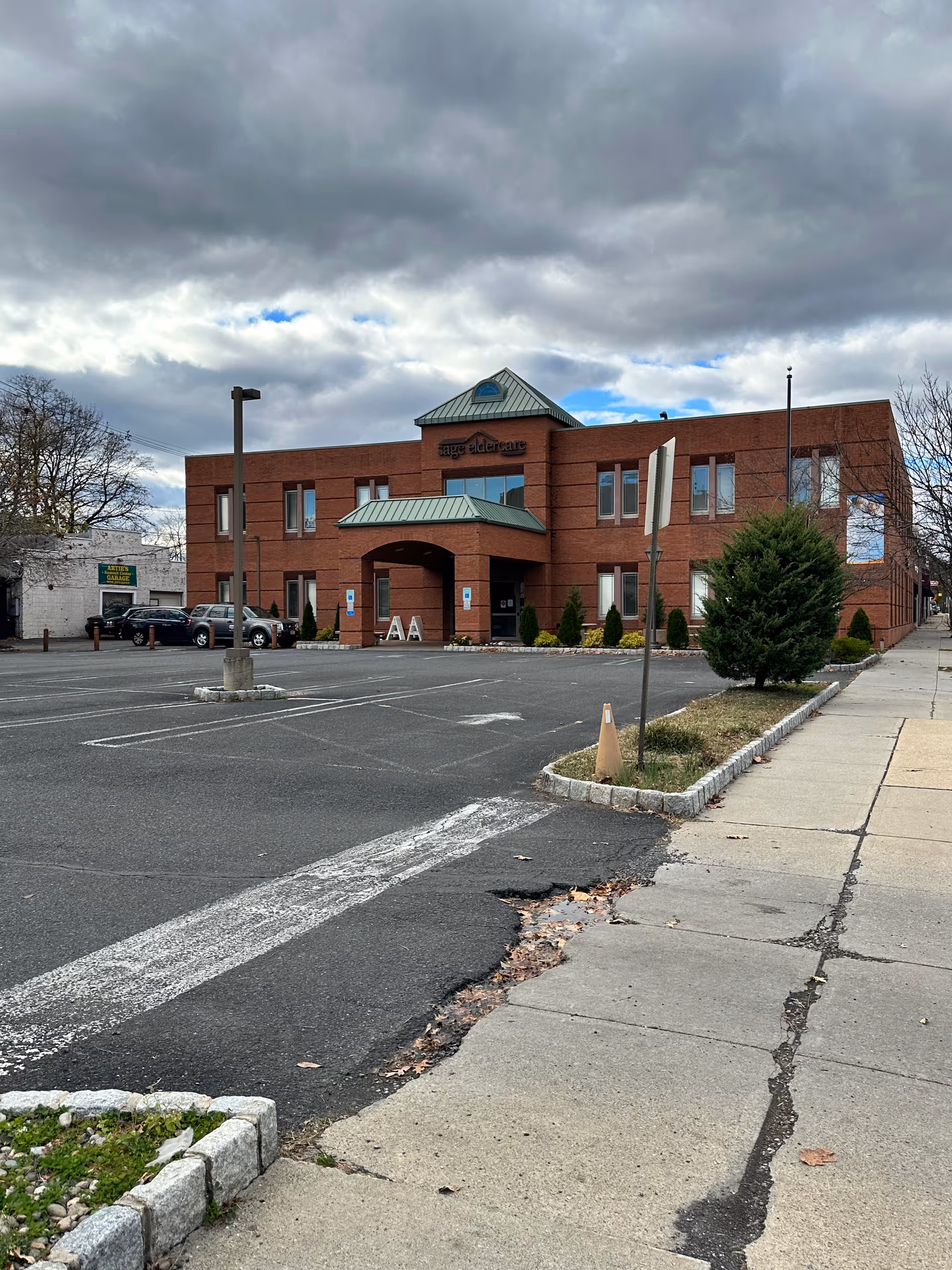 Red brick Sage Eldercare building with parking lot and walkway in foreground.