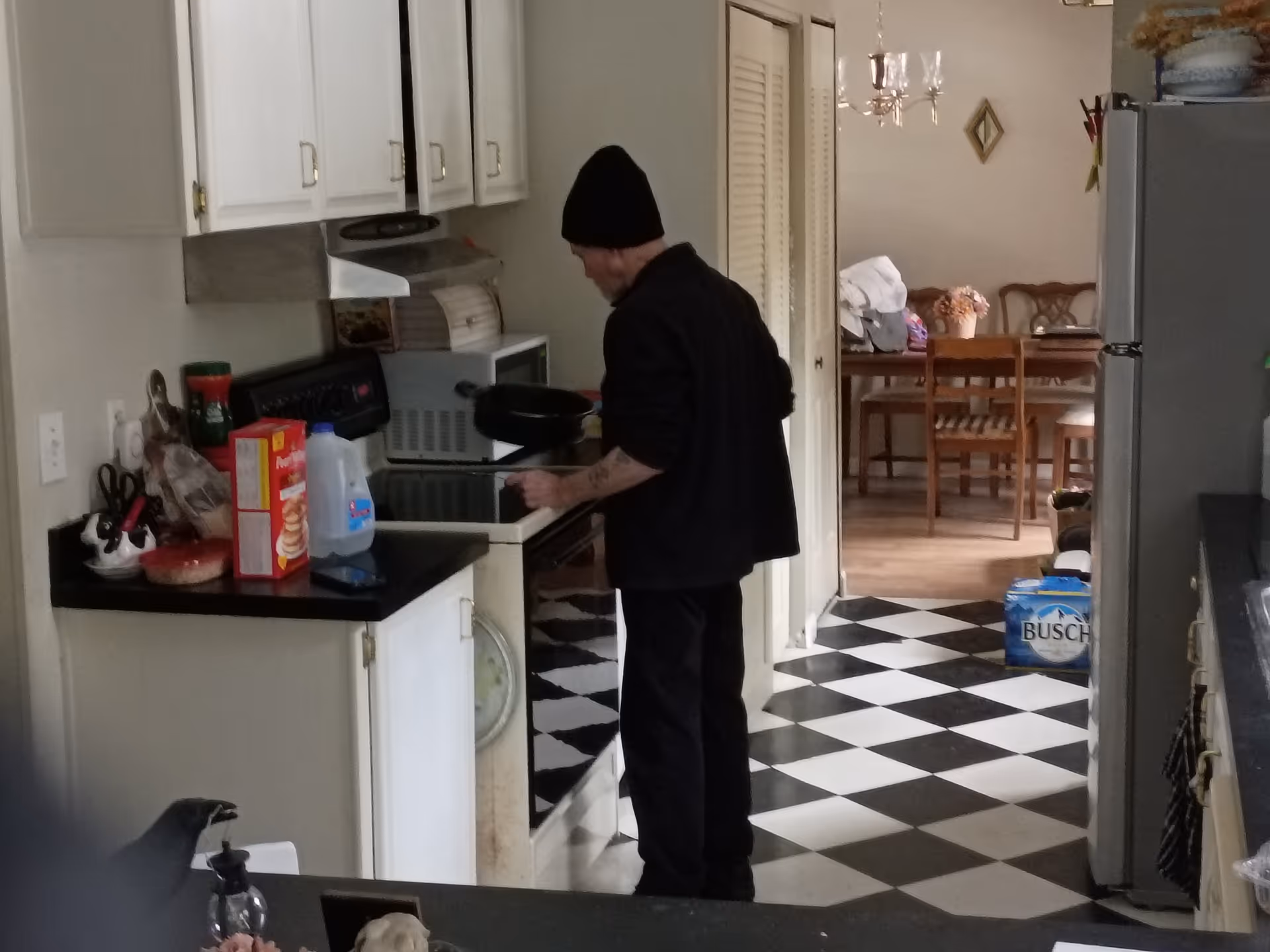 Man wearing a beanie cooks at a stove in a checkered black and white kitchen.