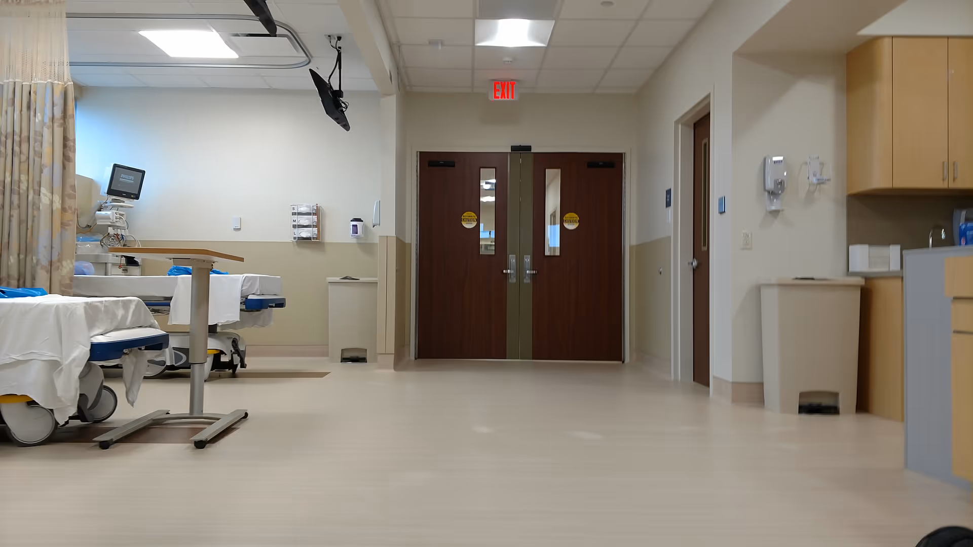 Hospital corridor with unoccupied beds, medical equipment, and double doors in the center.