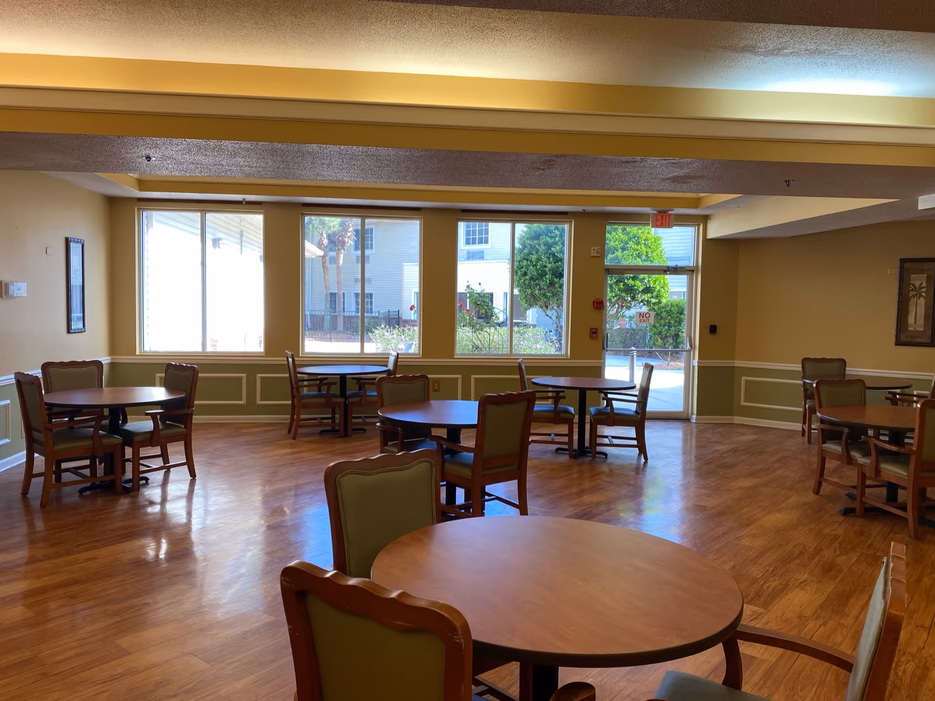 A dining room with several round wooden tables and green cushioned chairs arranged neatly on a wooden floor. Large windows and a glass door allow natural light to fill the room, showing an outdoor view with trees and buildings. The walls are painted in a warm beige color with white trim and decorative molding.