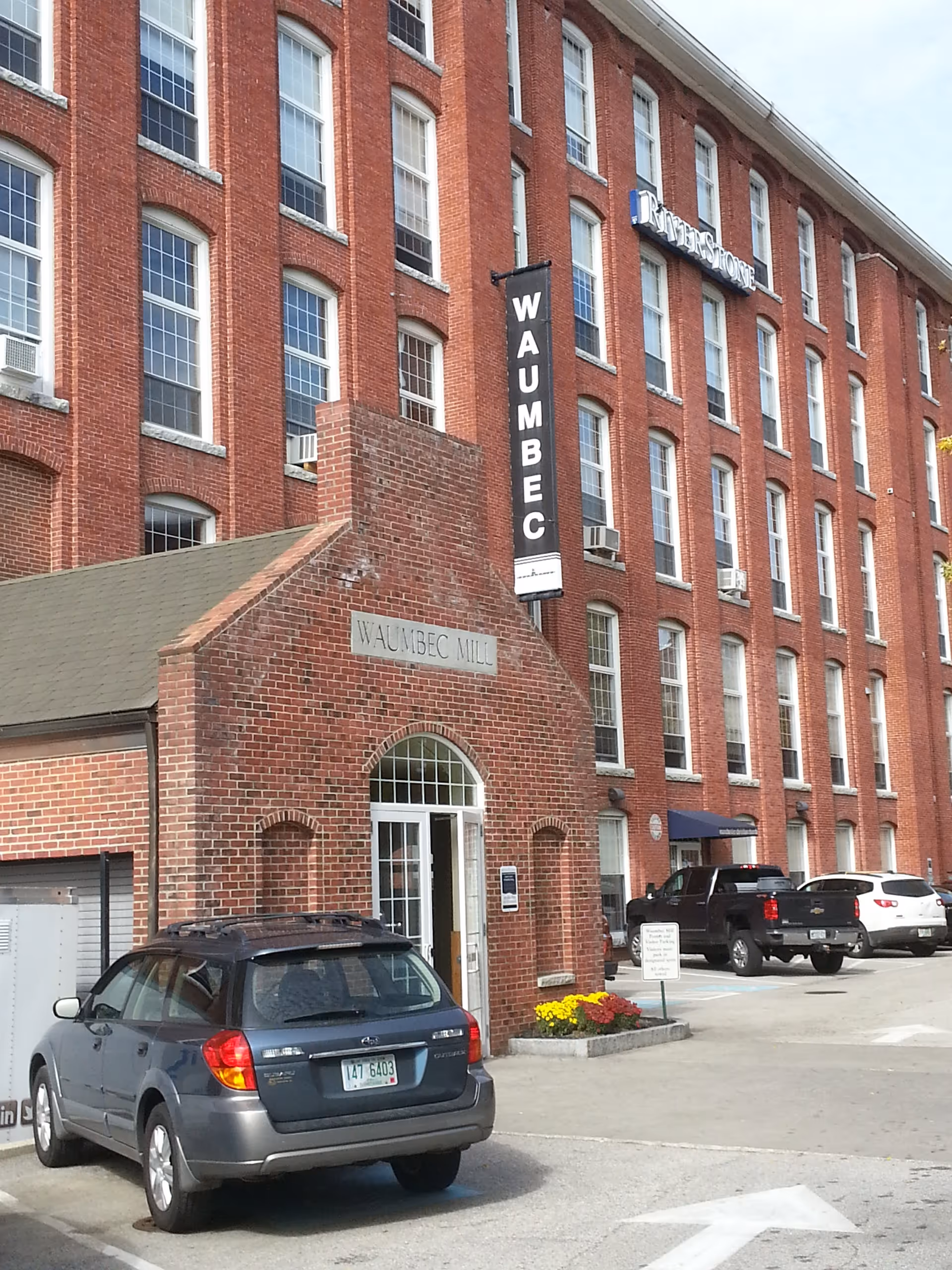 An exterior brick building with WAUMBEC MILL sign and parked cars.