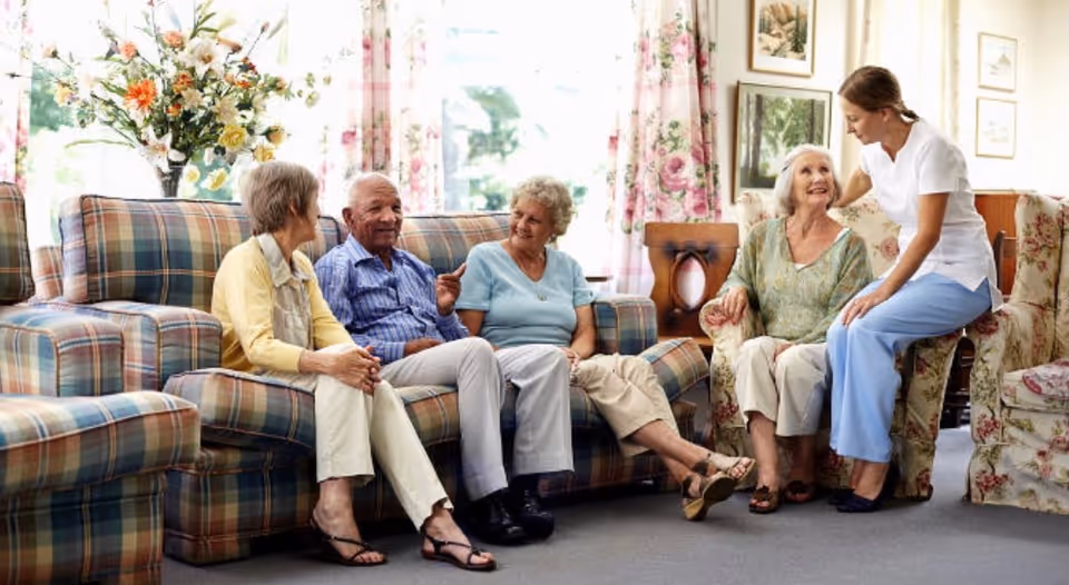 A group of elderly residents and a caregiver sitting and chatting in a bright living room with floral curtains and patterned sofas.