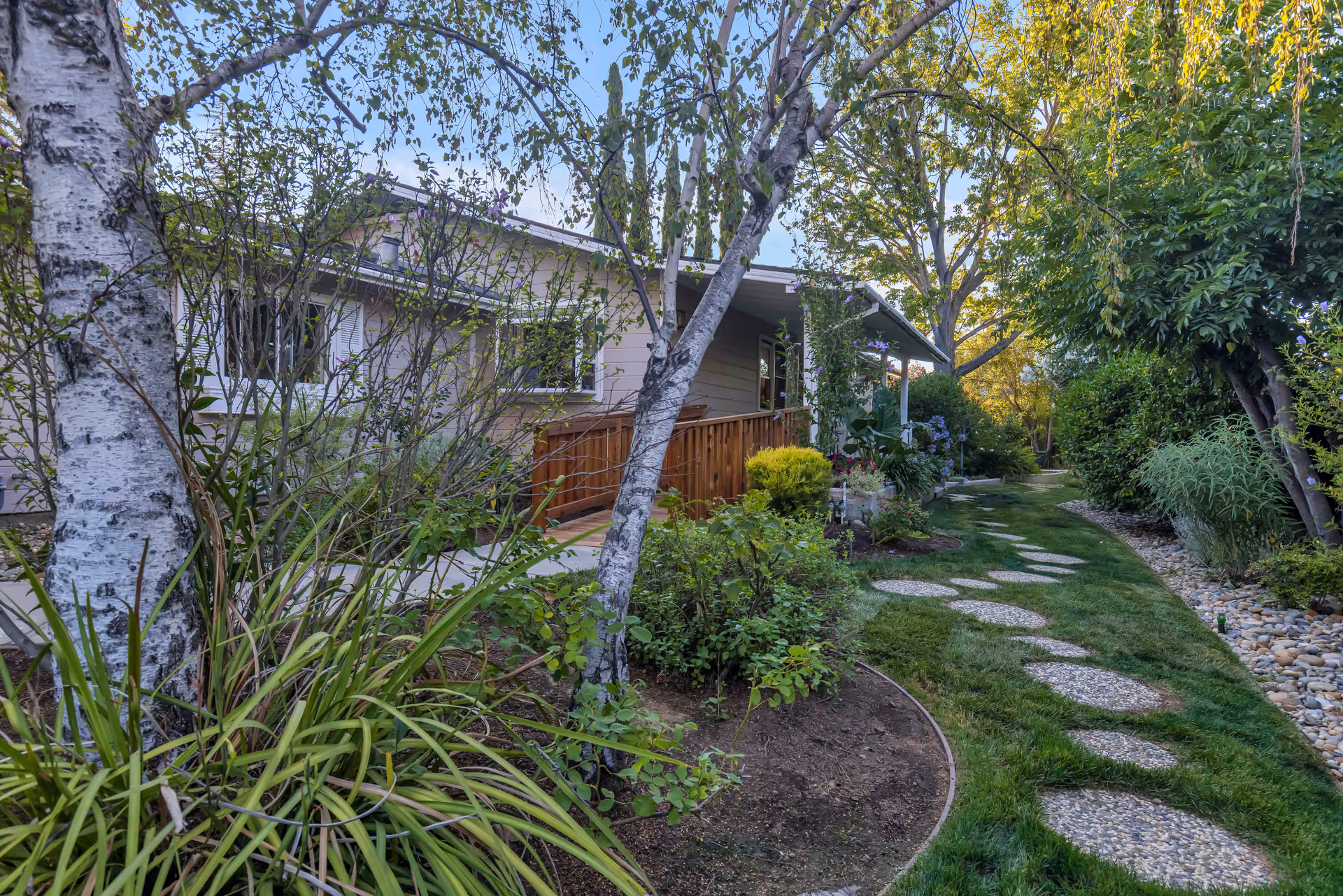 A garden path with circular stepping stones leading through a lush green yard with various trees, bushes, and plants. A single-story building with a wooden ramp and windows is visible on the left side, partially obscured by the foliage.