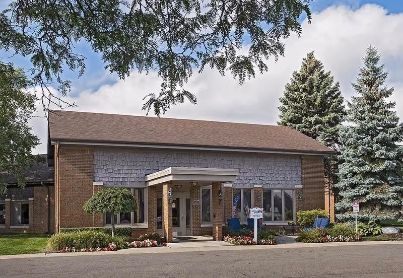 Front exterior of a single-story brick building with a covered entrance, landscaped flowerbeds, and tall evergreen trees.