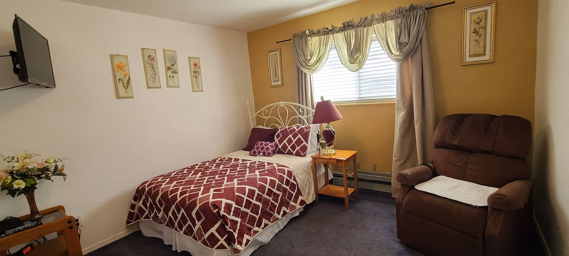 Cozy bedroom featuring a burgundy quilted bed, nightstand lamp, window with drapes, and a brown recliner.