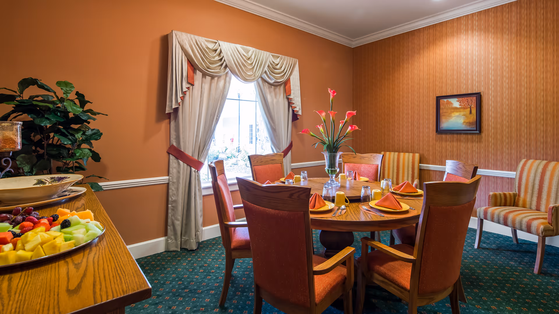 A dining room with a round wooden table set for six people with orange napkins and plates. There are six orange upholstered chairs around the table and two striped armchairs against the wall. A vase with pink flowers is in the center of the table. The room has orange walls, a window with cream and orange drapes, green carpet with a small pattern, and a sideboard with a bowl of fresh fruit and a plant.