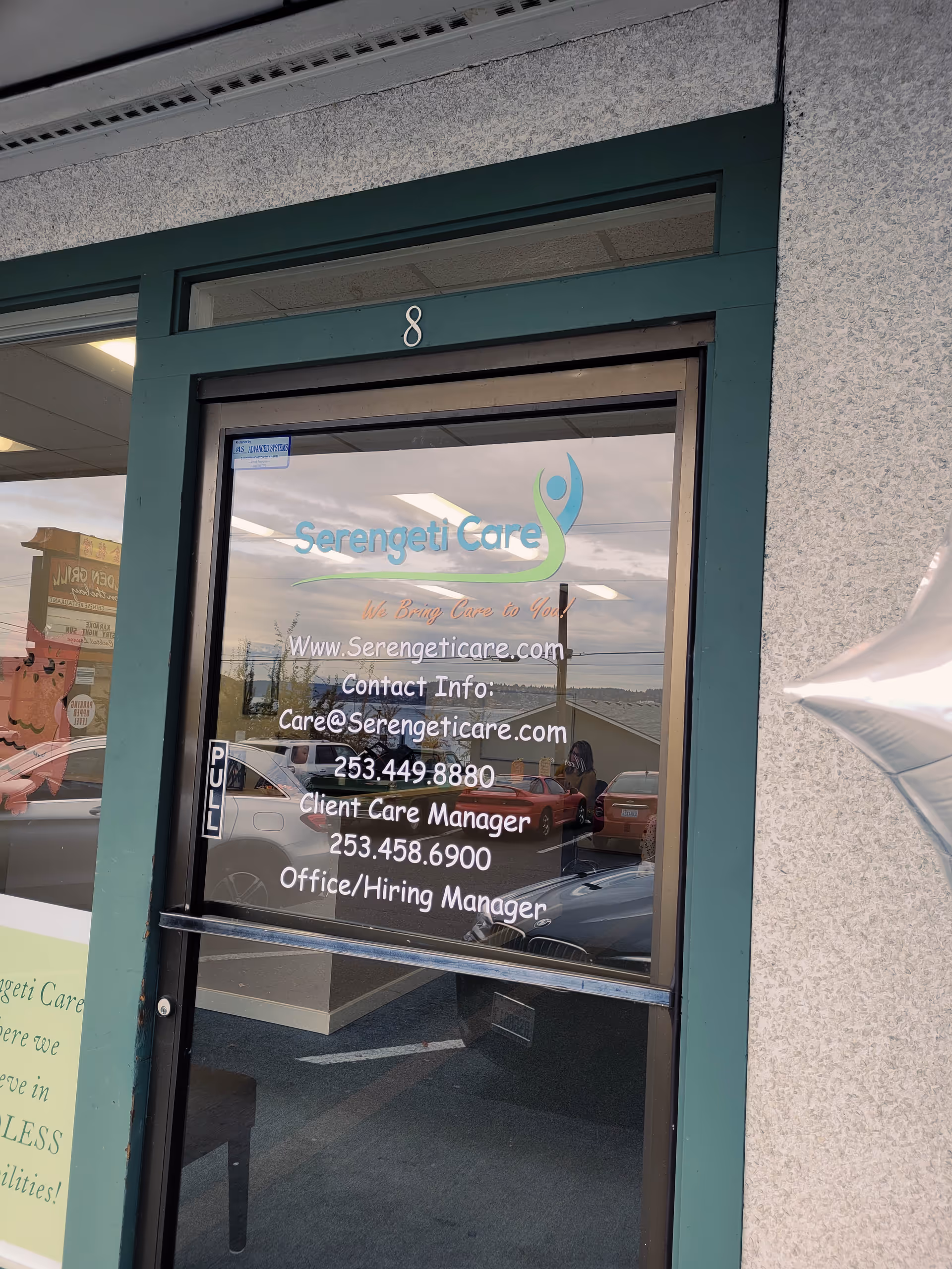 Glass door entrance of Serengeti Care with logo, contact info, and car reflections.