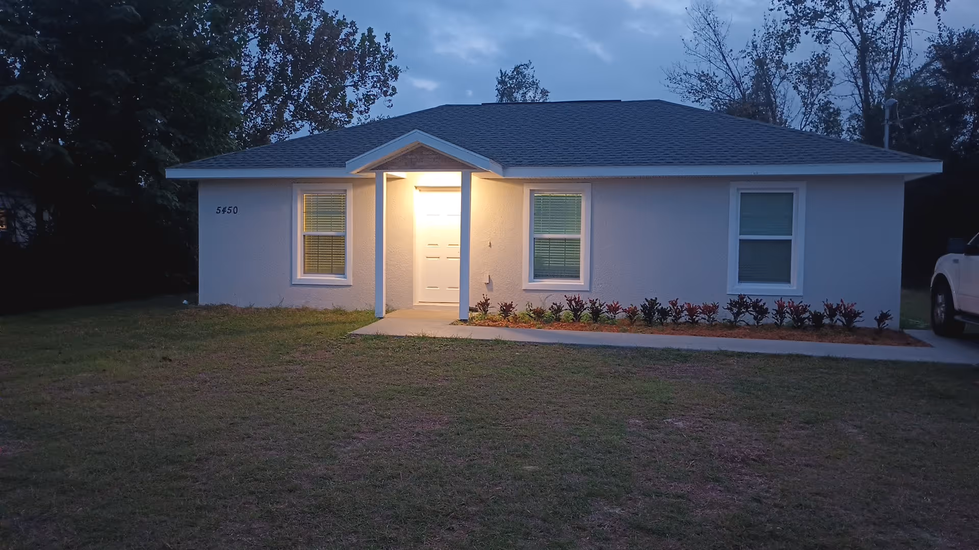 Single-story residential building with lit front entry, manicured shrub bed, and lawn at dusk.