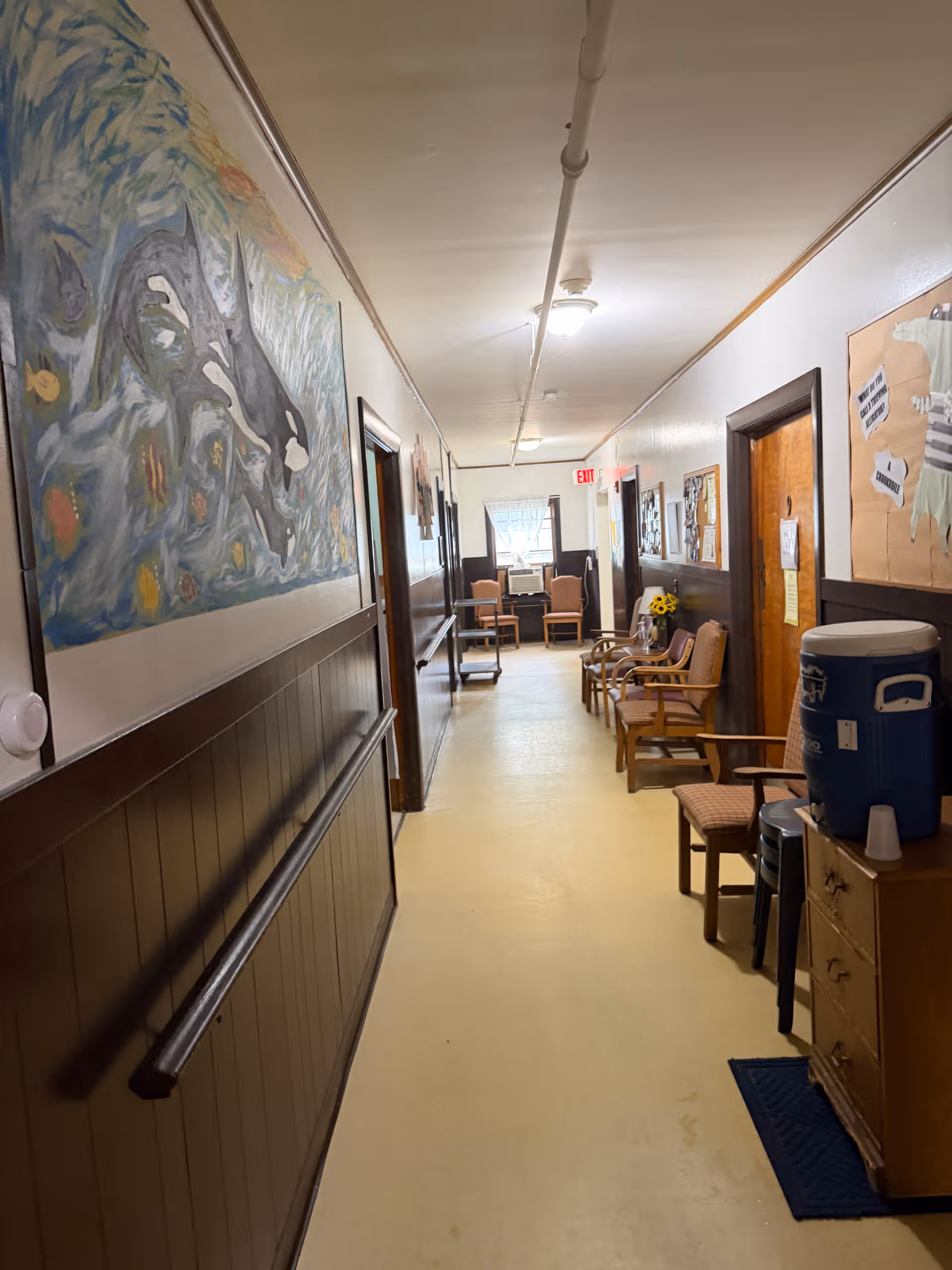 Narrow hallway in a senior living facility with chairs, artwork, and a water cooler.
