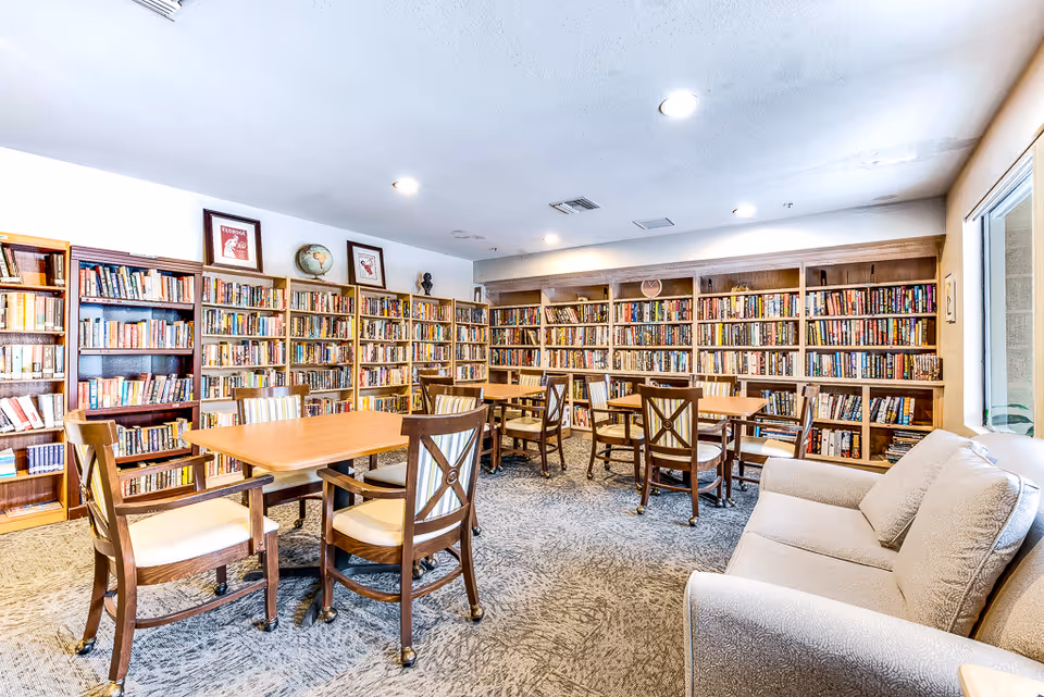 Bright senior living library with long bookshelves along walls, tables, chairs, and a beige sofa.